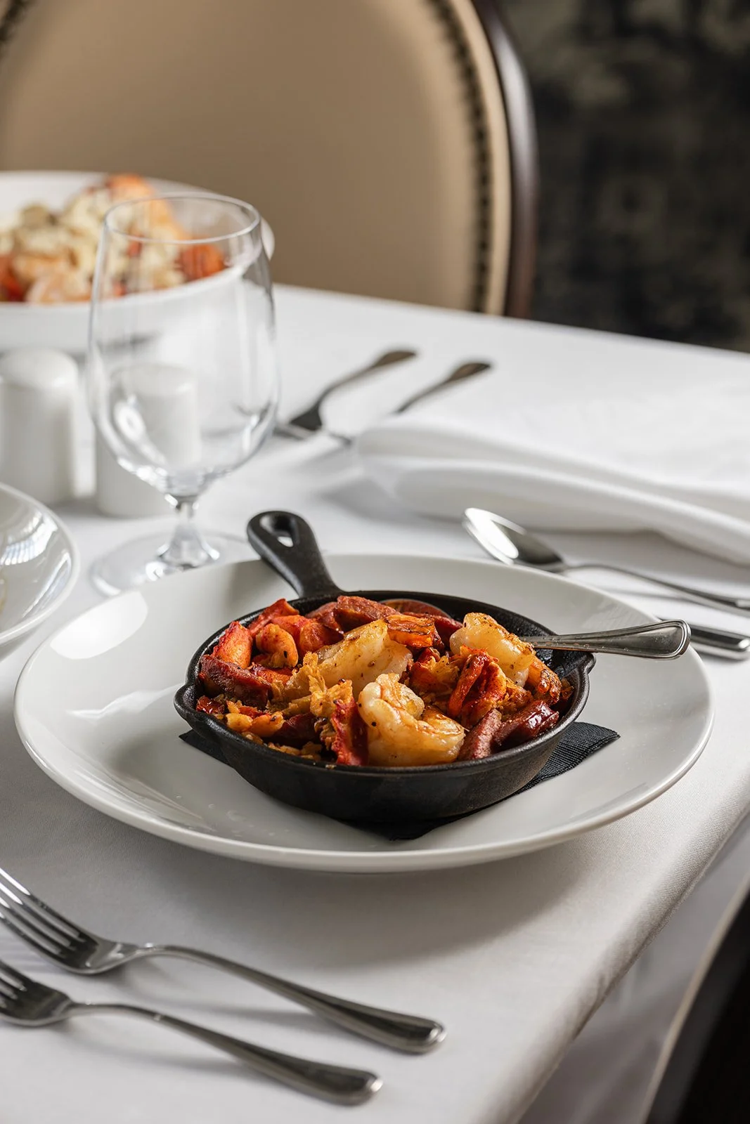 A table setting with a small cast iron skillet of Southern Seafood Paella on a white plate. There is an empty wine glass, utensils, a white napkin, and a bowl of salad in the background.