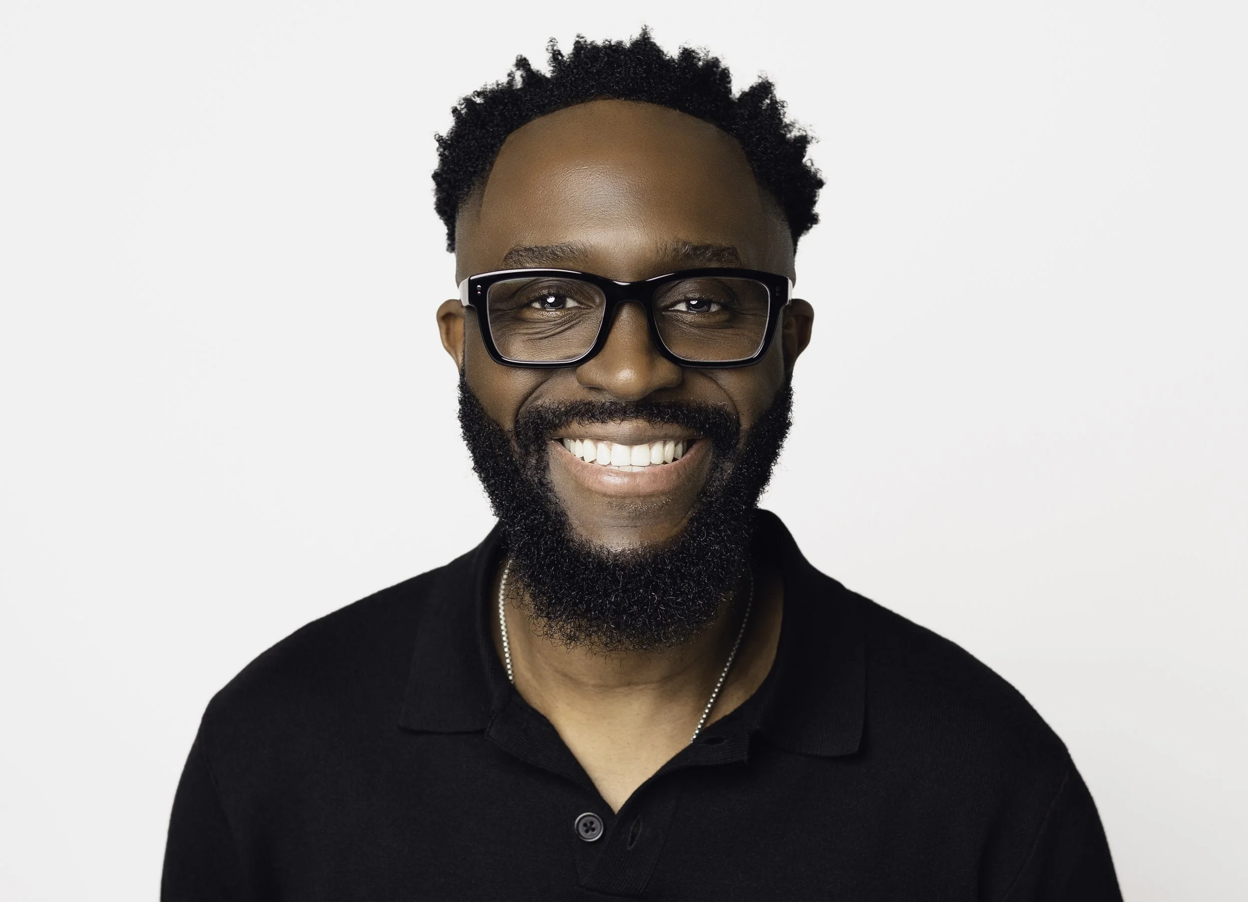 A smiling man with black hair, beard, and glasses wearing a black polo shirt against a white background.