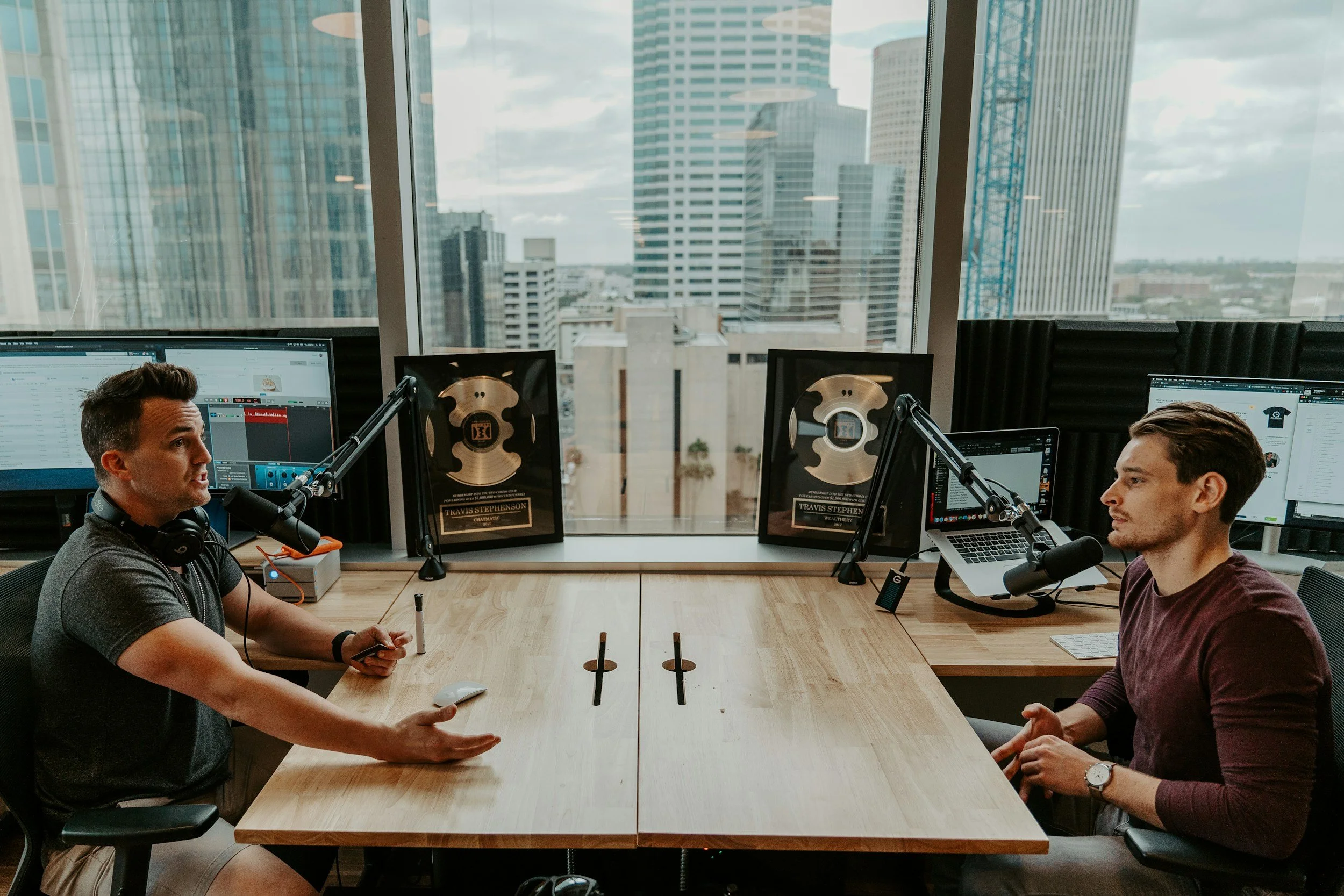 Two men having a conversation during a podcast recording session in a modern office with large windows showing a cityscape.