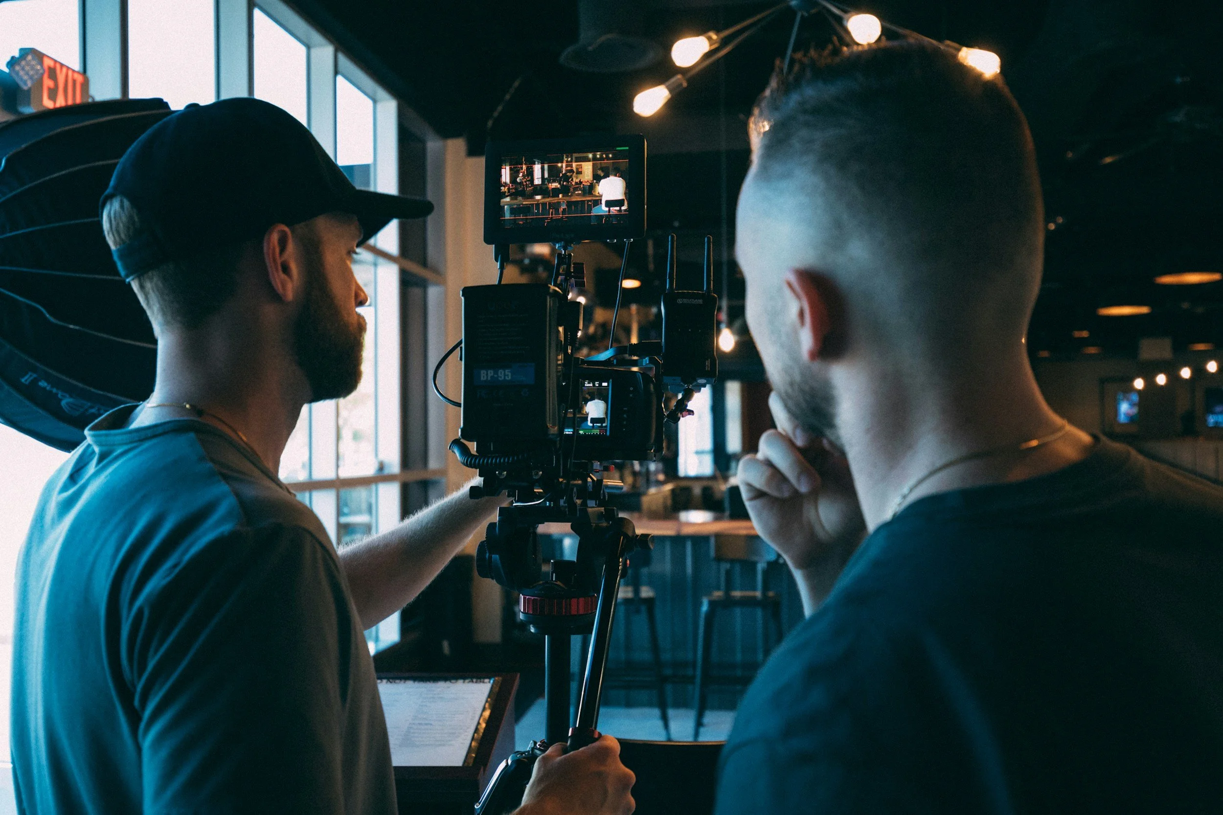 Two men on a film set: one adjusting camera equipment, the other observing with his hand on his chin, inside a dimly lit venue with large windows and hanging lights.