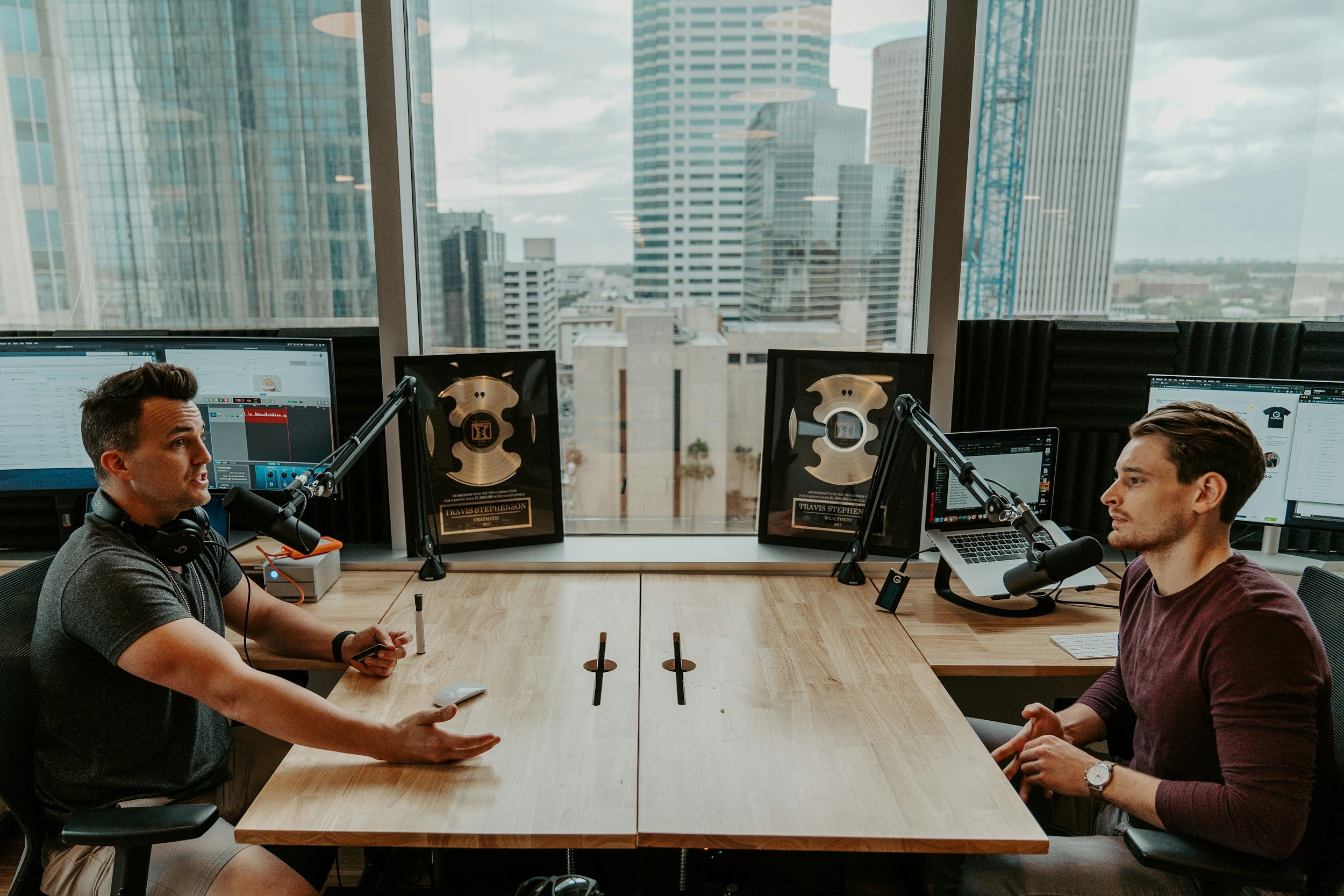 Two men sit at a conference table with podcasting microphones, headphones, and computer monitors, inside a high-rise office with large windows overlooking a cityscape.