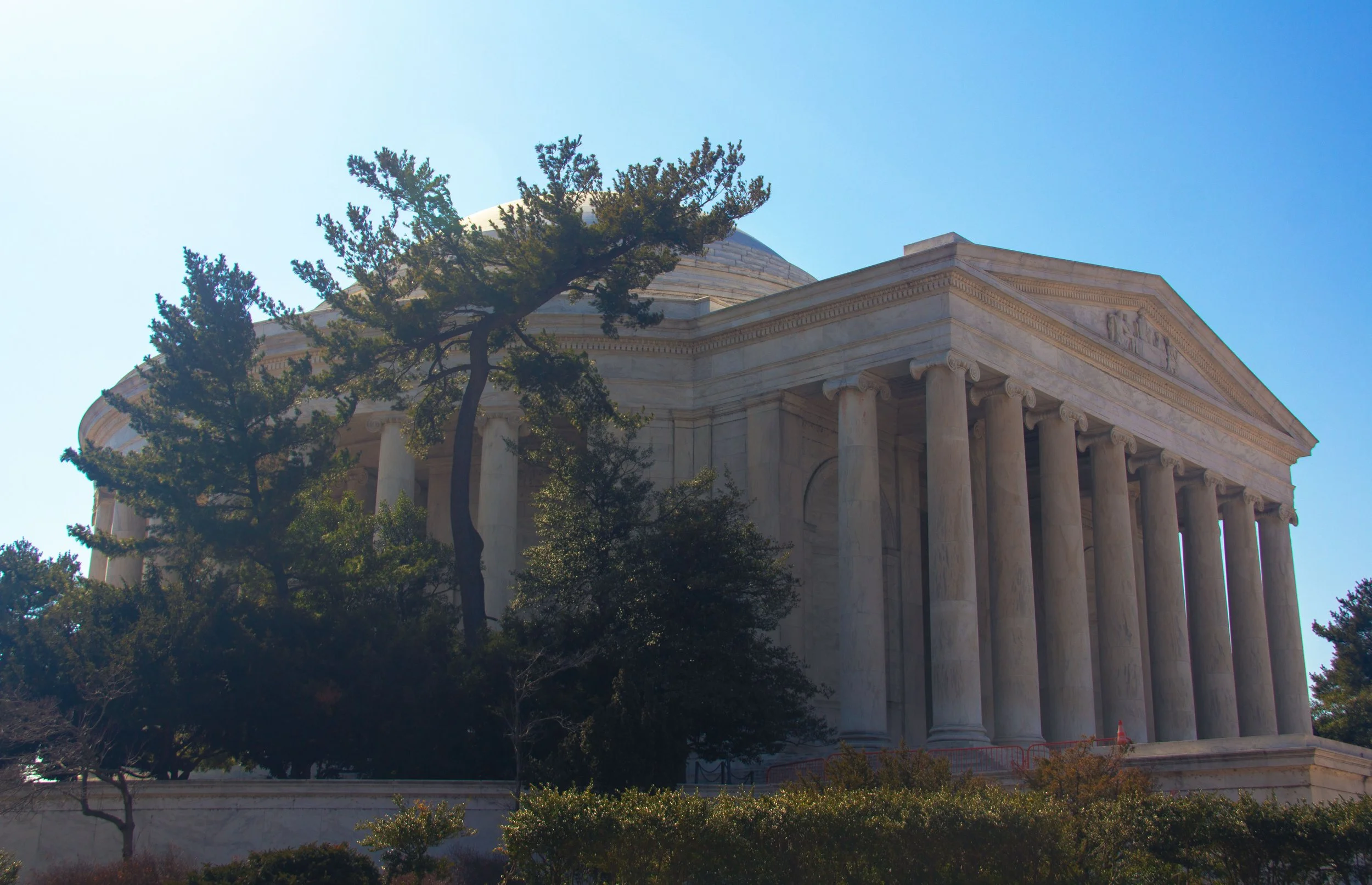 The Lincoln Memorial, a white marble monument with large columns, partially obscured by trees, against a blue sky.