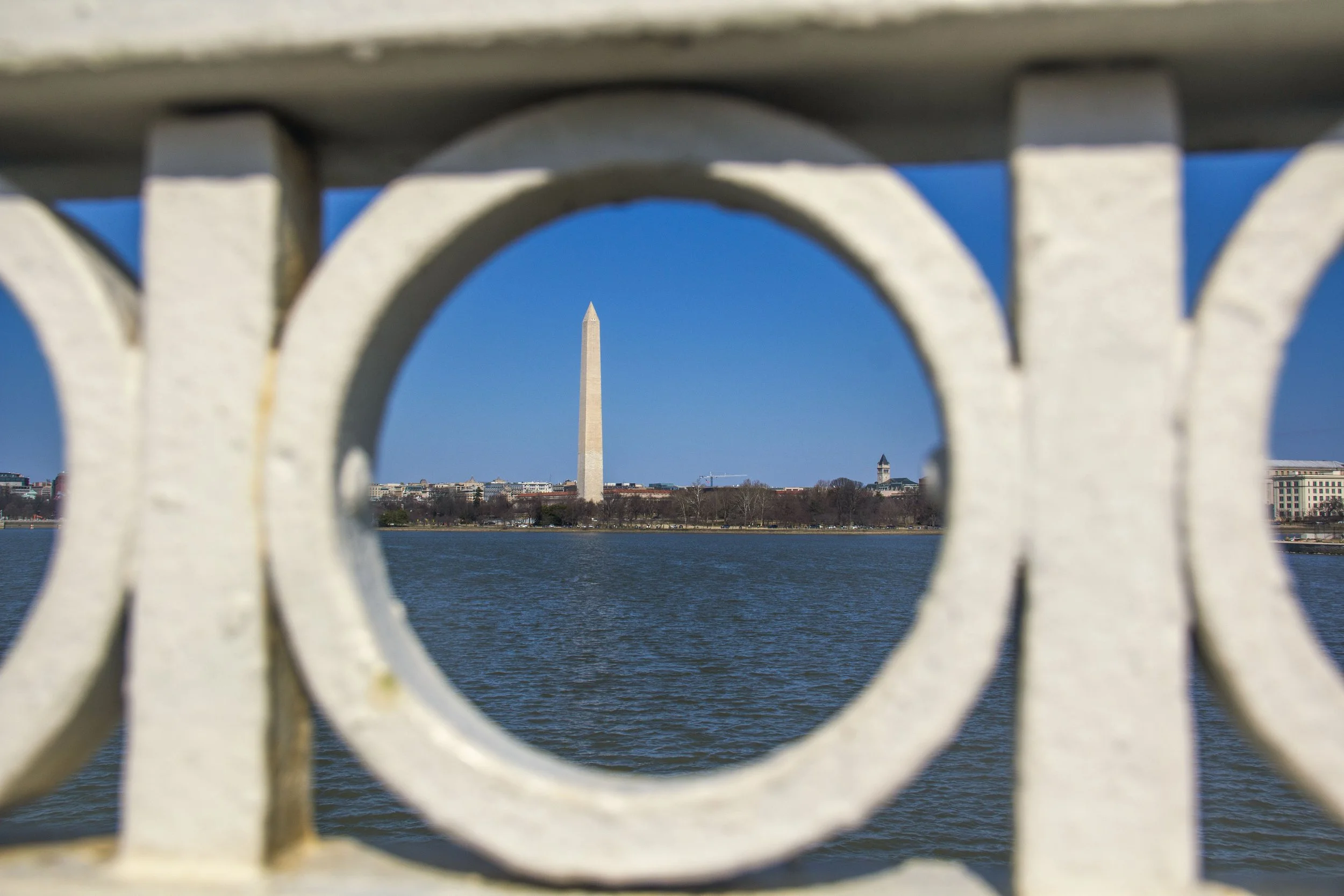 The image shows the Washington Monument in Washington, D.C., seen through the gaps of a white decorative iron fence along a body of water.