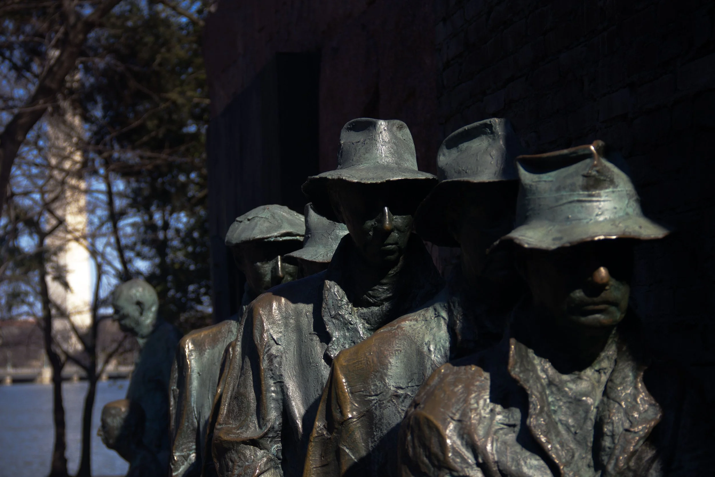 A bronze sculpture of a group of people, with several wearing hats, set against a brick wall and trees in the background.