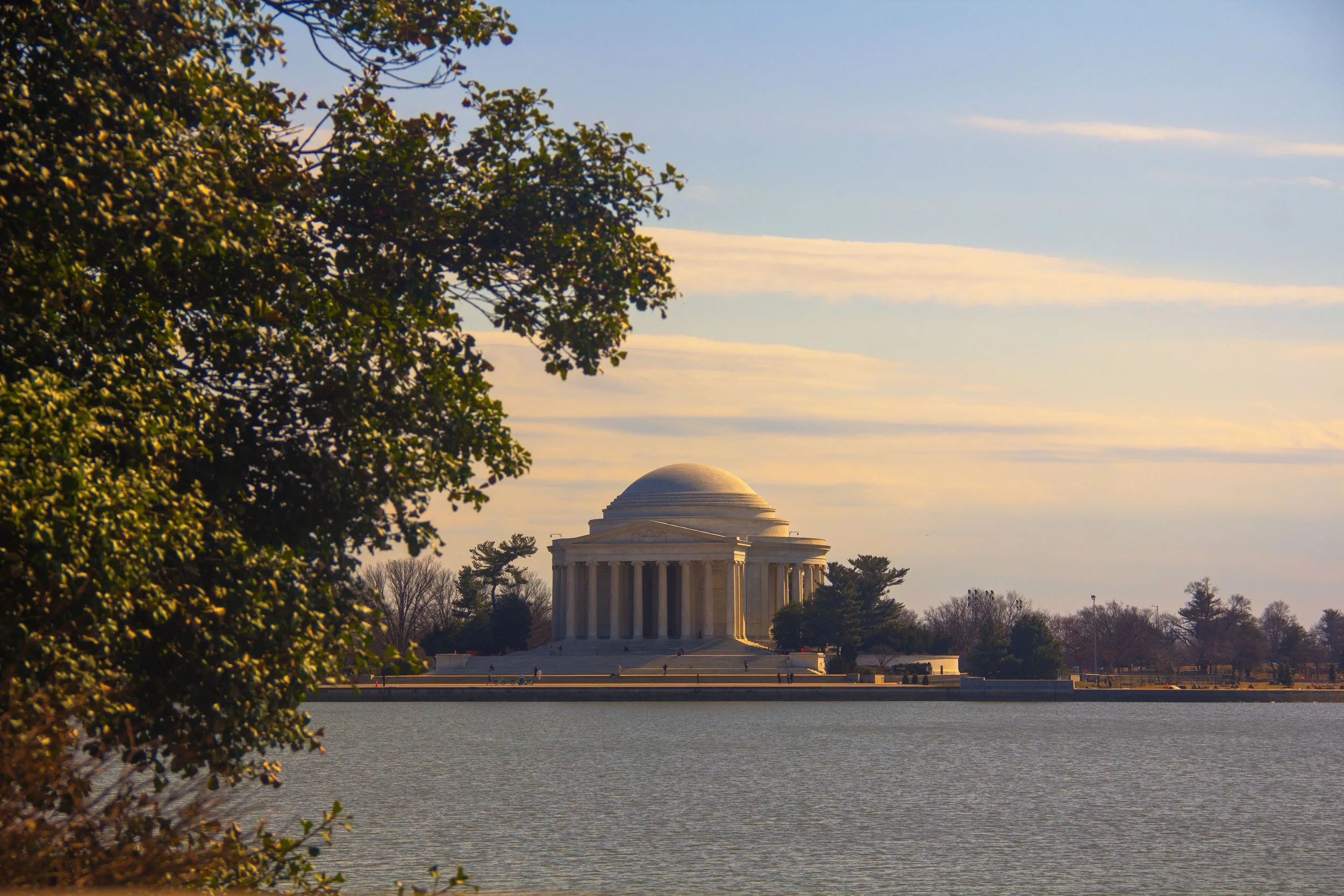 The Lincoln Memorial seen across a body of water with trees on the foreground and a sky with scattered clouds at sunset.