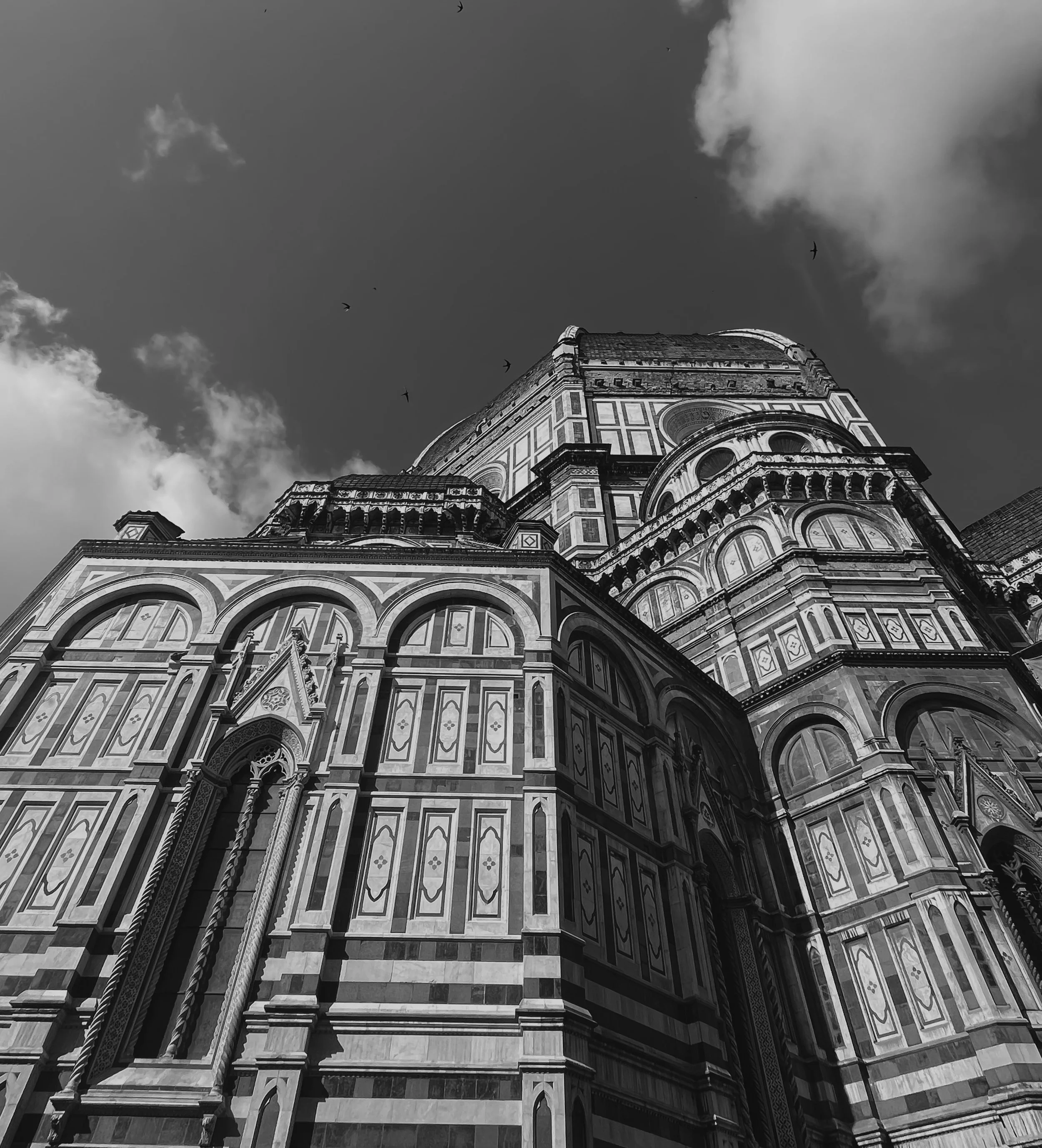 Black and white photo of the Florence Cathedral's dome, showcasing intricate architectural details and stonework, with a partly cloudy sky above and birds flying in the background.
