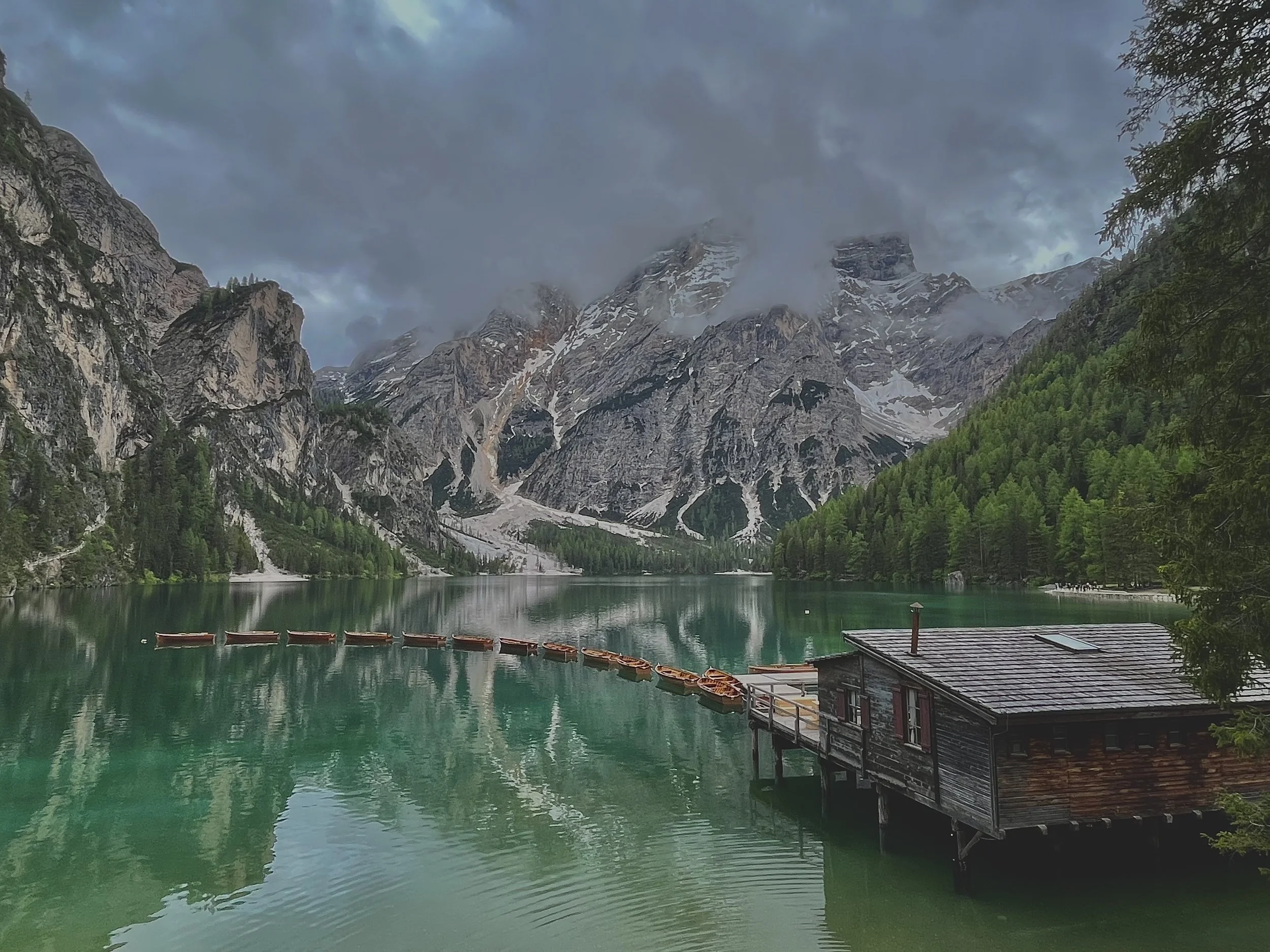 A scenic mountain landscape with a cloudy sky, a turquoise lake in the foreground, and snow-capped peaks in the background. There are boats on the lake and a wooden building on the shore.