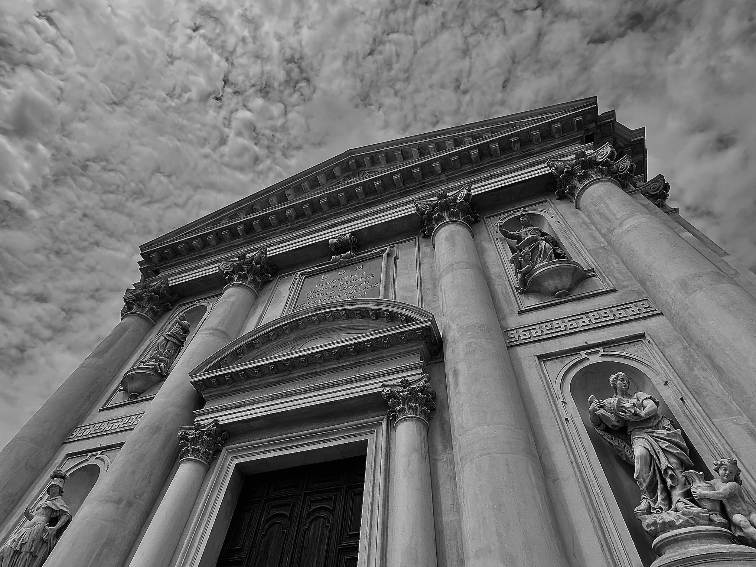 Black and white photo of the upper facade of a historic building with classical architecture. The building features large columns, ornate statues, and detailed cornices, set against a cloudy sky.