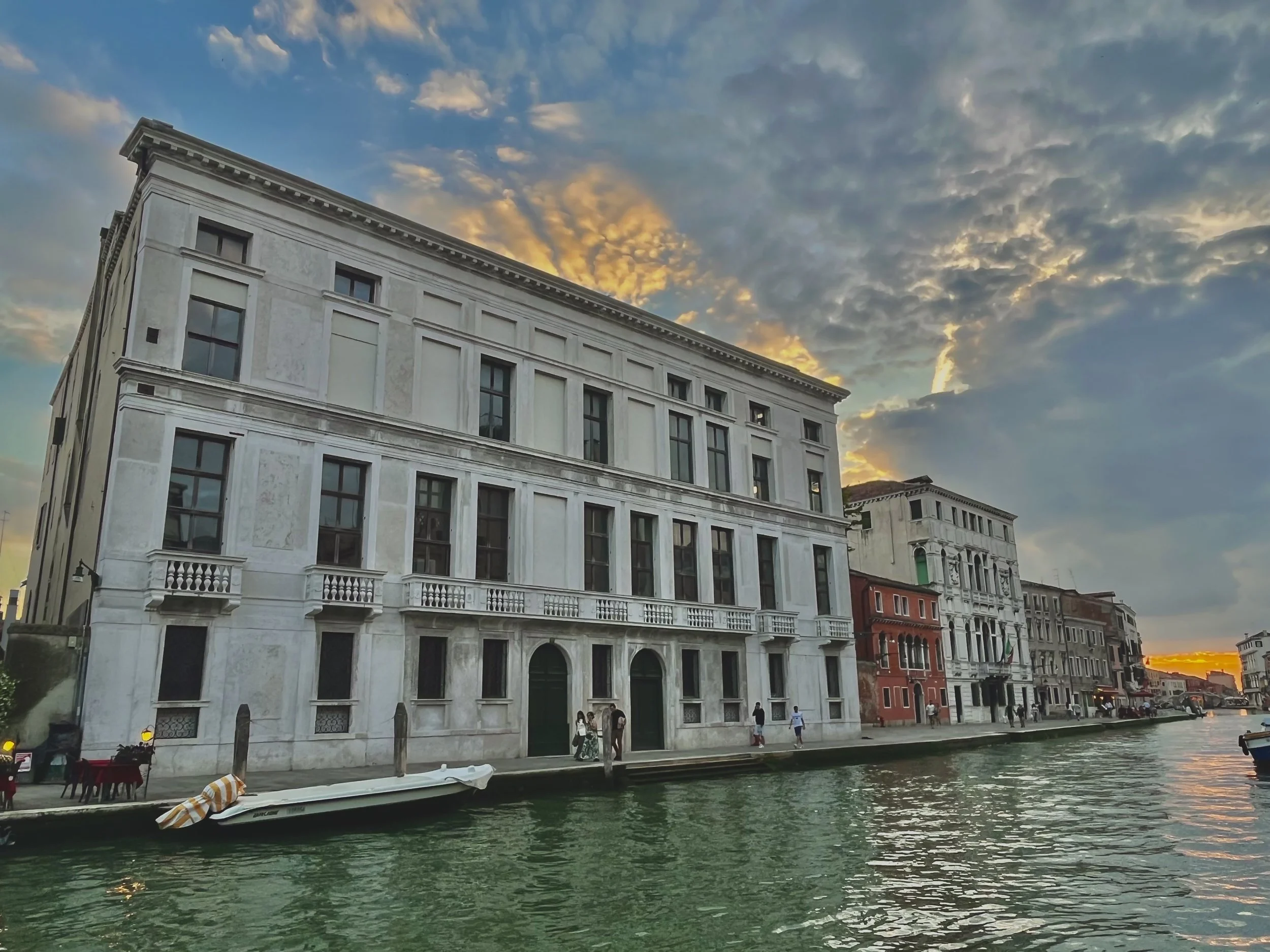 A cityscape with historic buildings along a canal at sunset, with people walking along the water's edge and the sky filled with clouds and a warm glow from the setting sun.