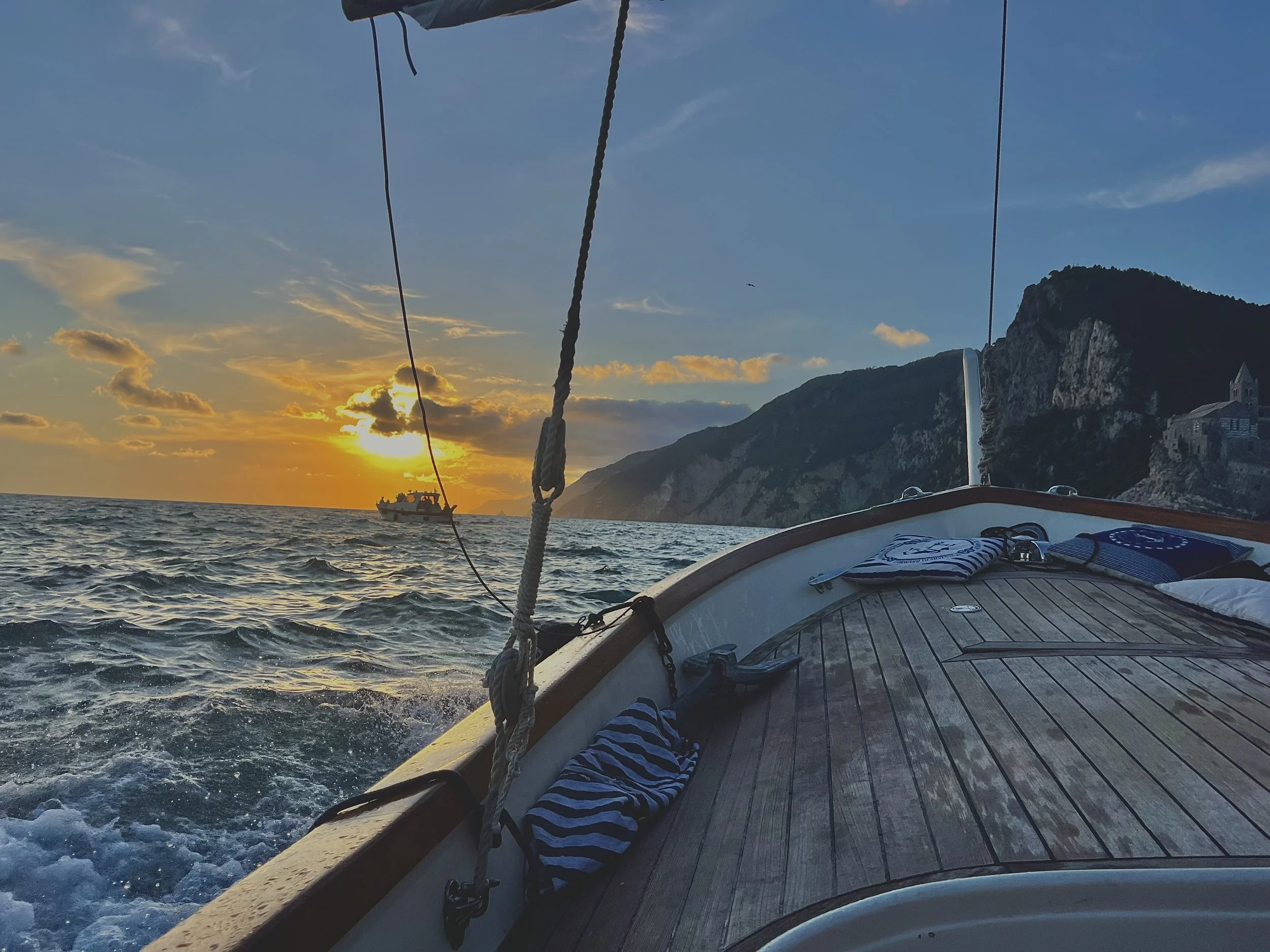 View from a boat at sunset showing the deck and railing in the foreground, the ocean with gentle waves, a distant boat, and a mountainous coastline with a church on the right, under a sky with scattered clouds.