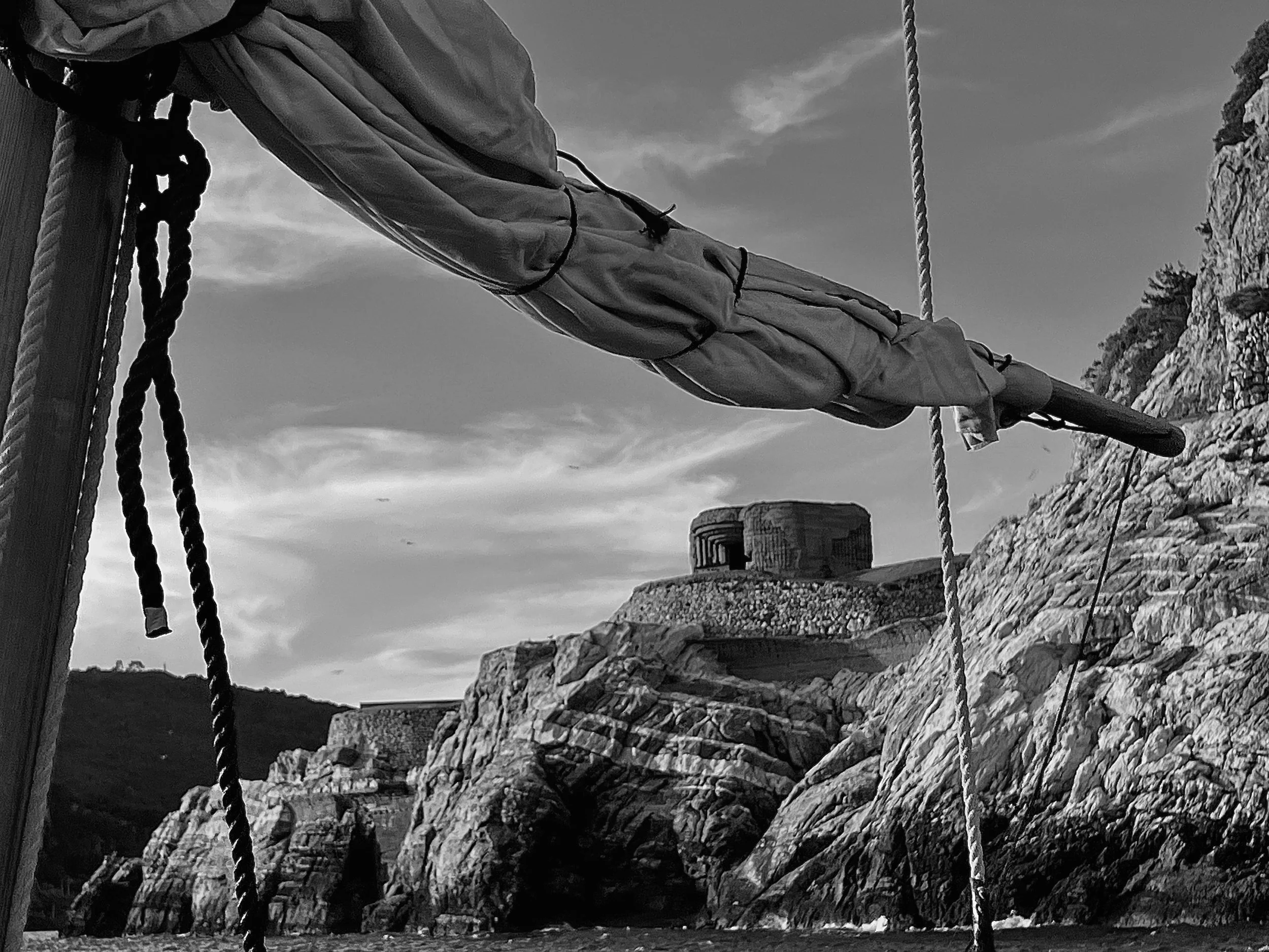 Close-up view of a sailboat's mast with a rolled-up sail secured by ropes, set against a rocky hillside and cloudy sky in black and white.