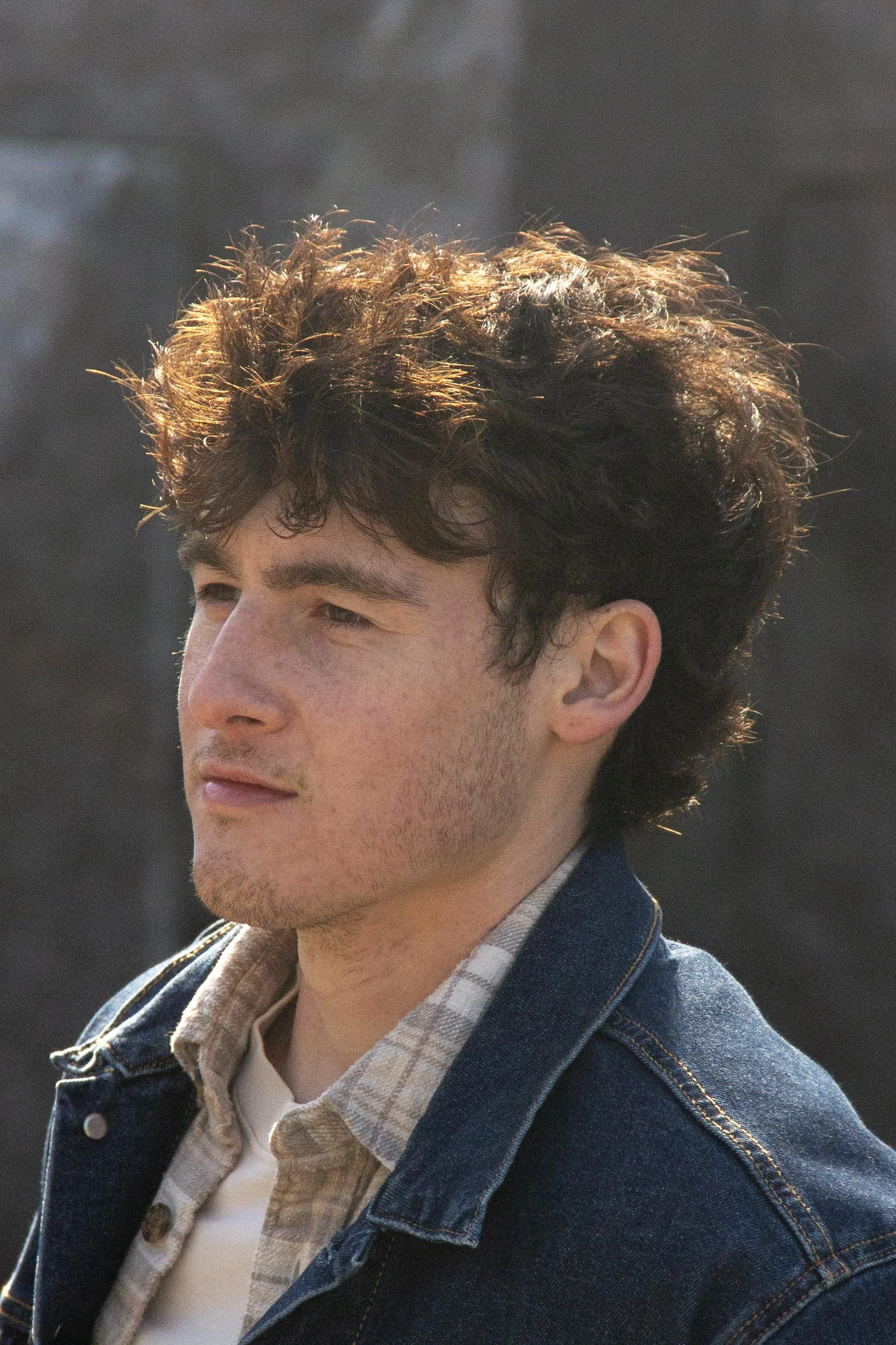 Close-up of a young man with curly brown hair, wearing a plaid shirt and denim jacket, with a blurred background.
