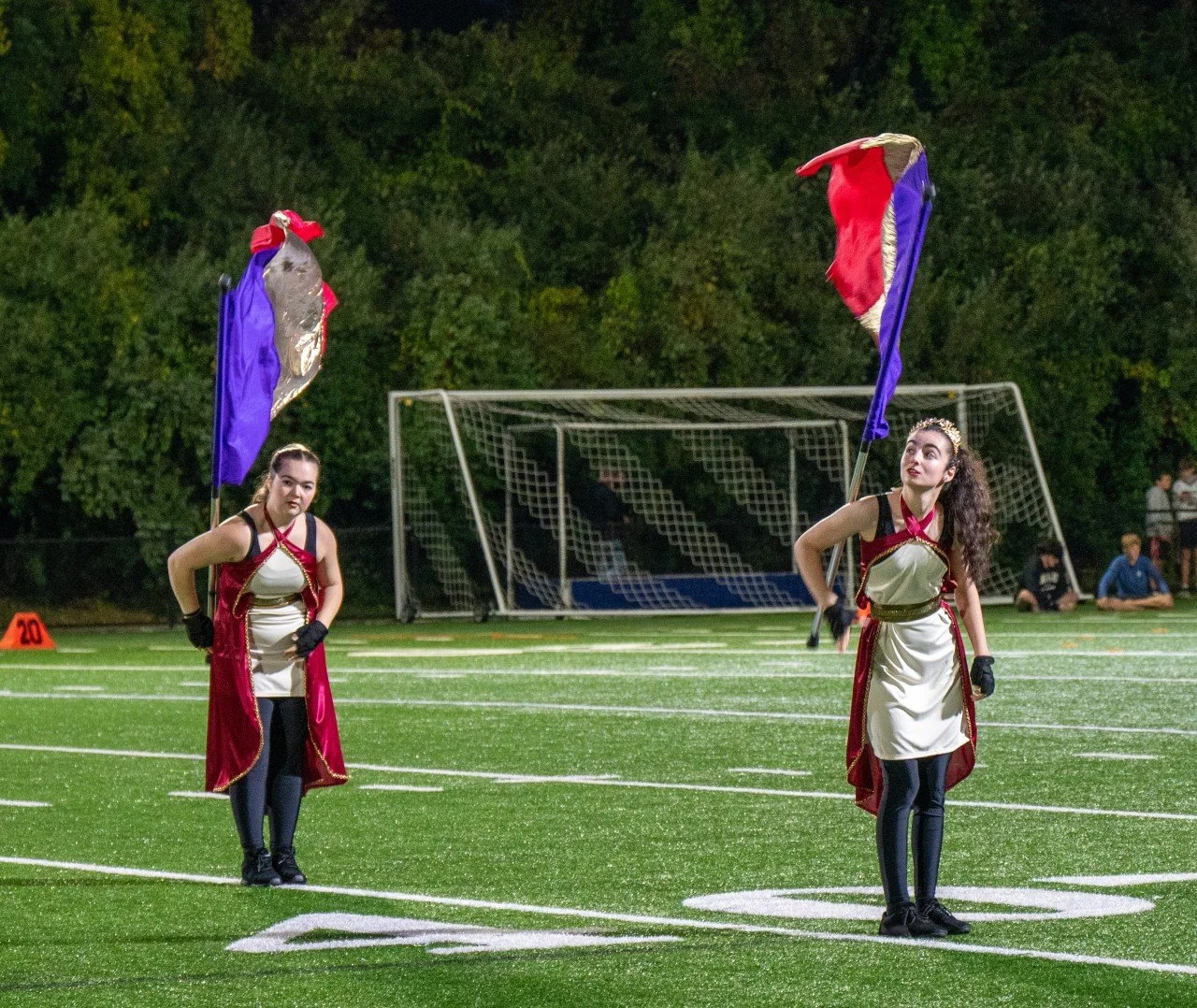 Two young women dressed in spirit uniforms holding large flags on a football field at night, with a goalpost and trees in the background.