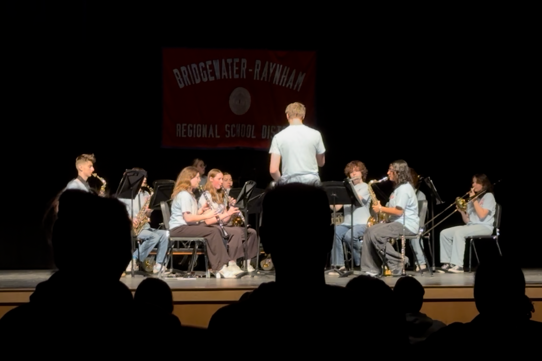 Youth orchestra performing on stage at a school district event, with players seated in two rows, and a conductor standing in front facing them. A banner with 'Bridgwater-Raynham Regional School District' hangs in the background.
