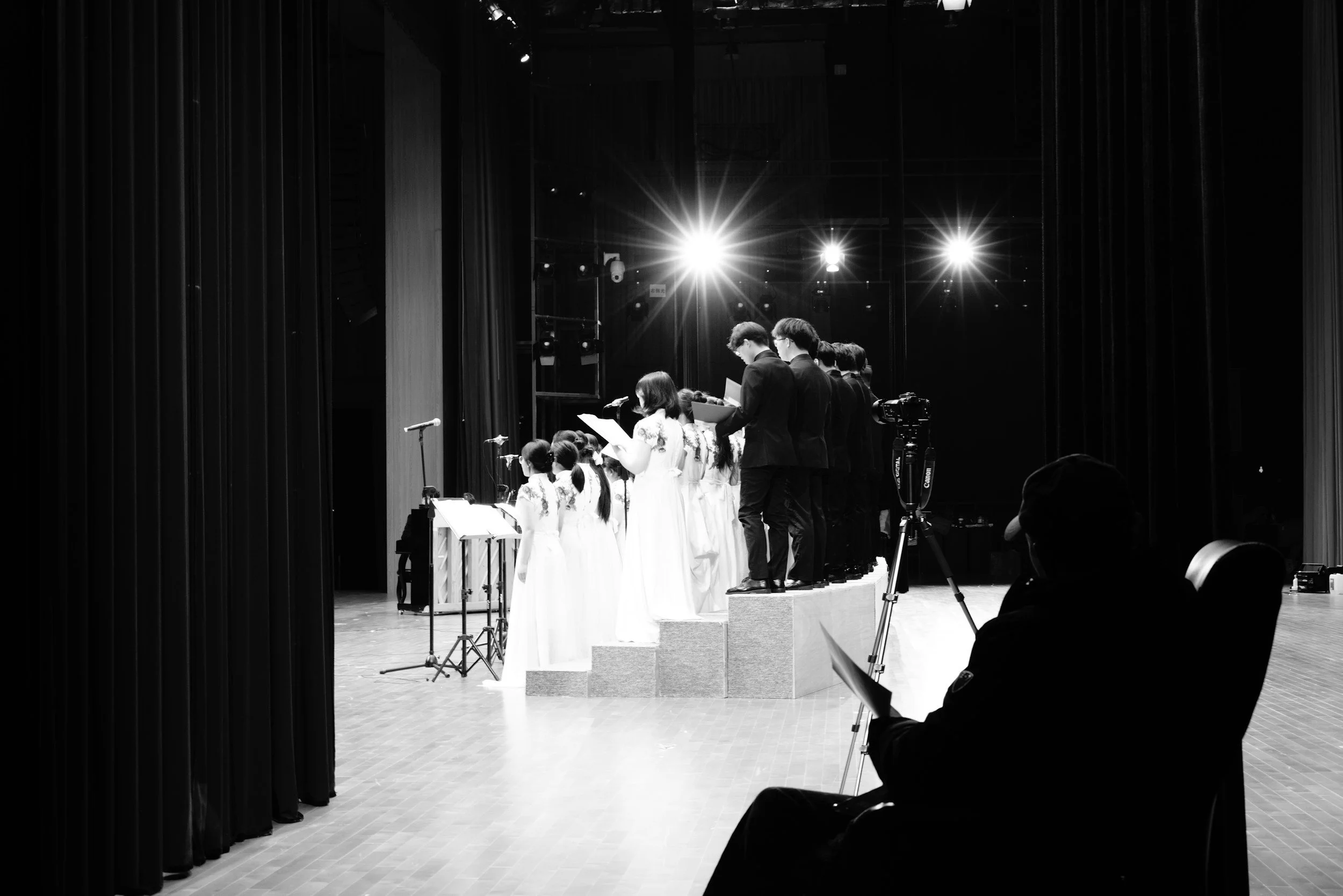 A group of children and adults performing on stage, possibly singing, with stage lights shining behind them in a theater or auditorium setting.