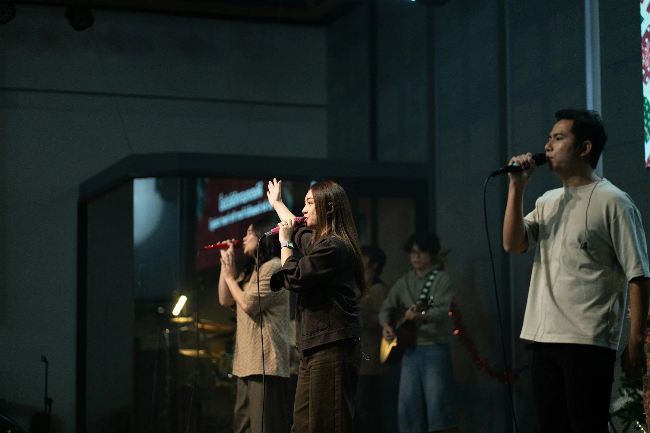 Group of people singing into microphones on stage in a dark room, with a large screen displaying text behind them.