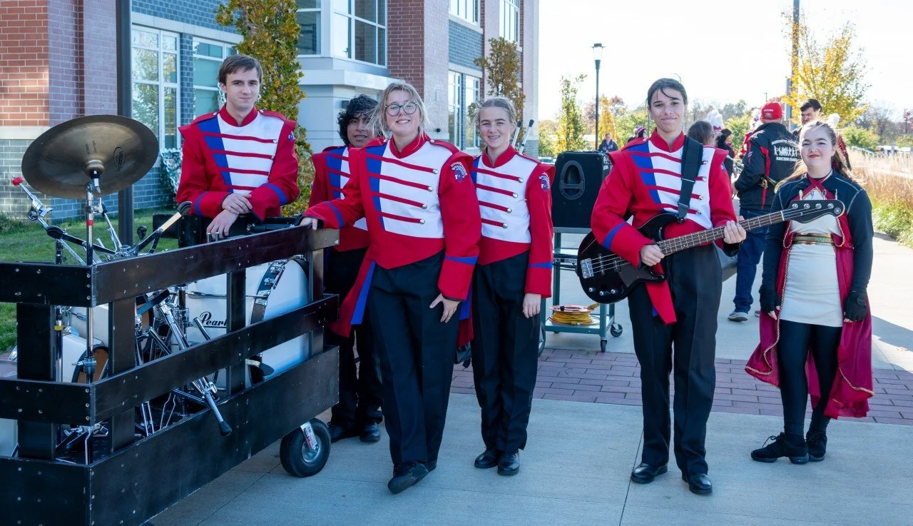 Group of young band members in marching band uniforms standing outdoors, with musical instruments including a drum set and an electric guitar, in front of a modern brick building and trees with autumn leaves.