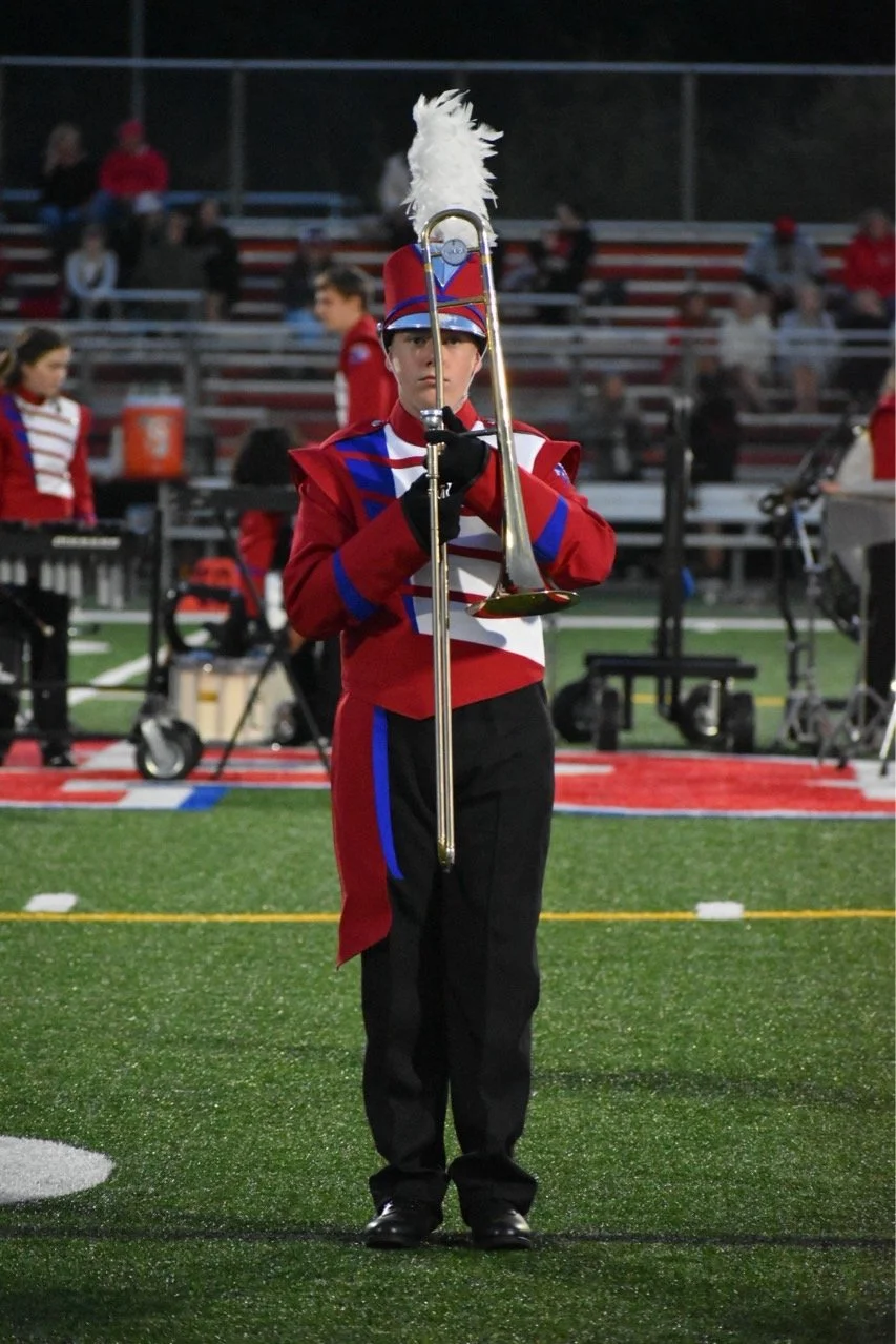 A young marching band member in a red, white, and blue uniform with a feathered hat, standing on a football field and holding a brass instrument upright.