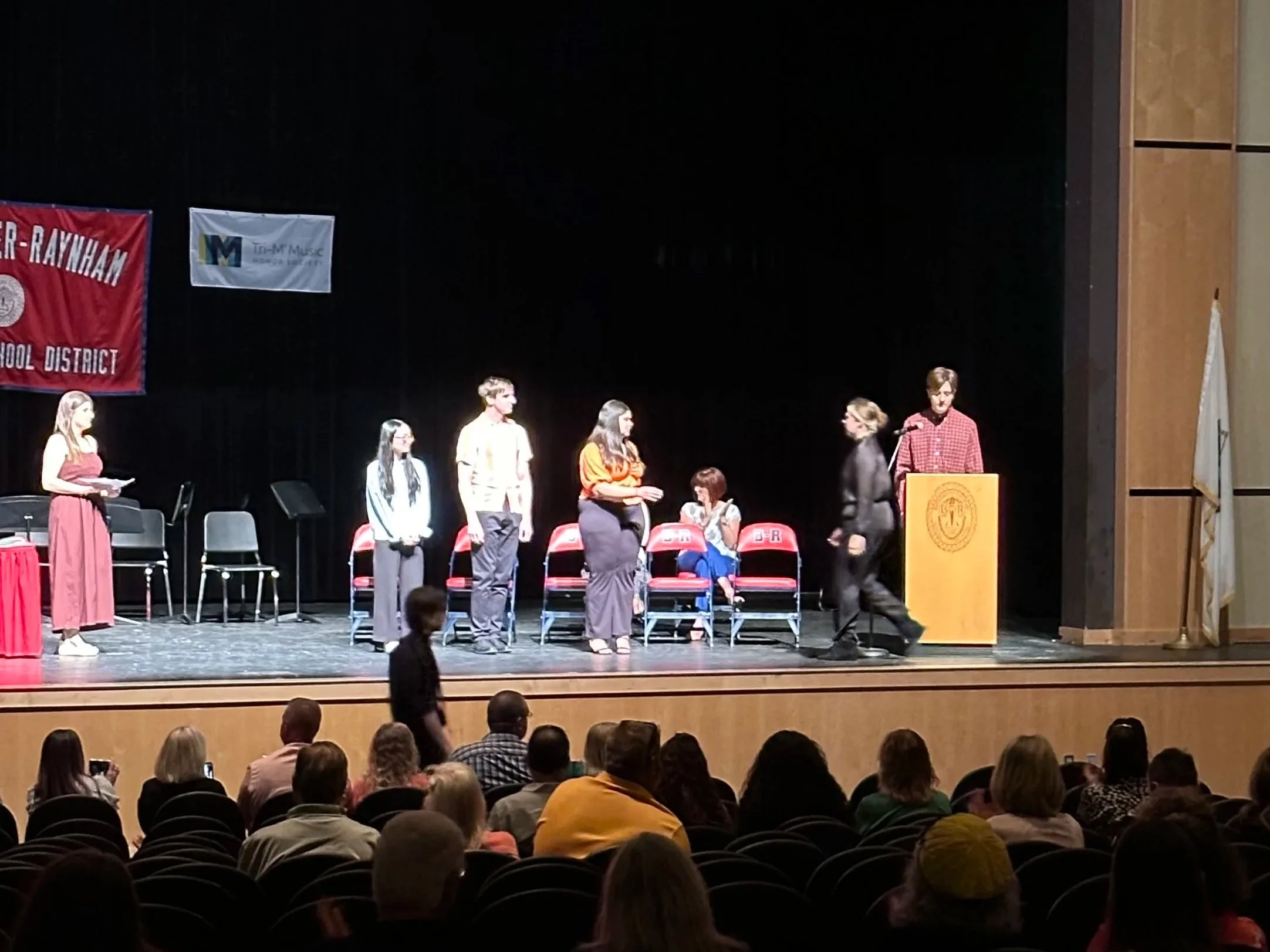 People gathered in an auditorium with a stage, some standing and others seated. There are banners on the stage, one says 'R-RAYNHAM' and another has a logo for 'Tu-M Music'. The audience is watching the event attentively.