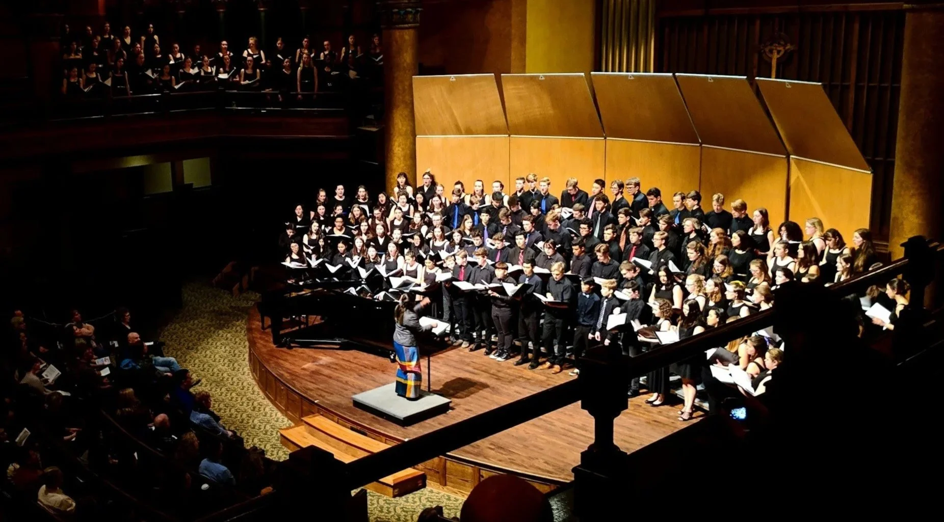 A large choir of young people singing on stage in a concert hall, directed by a conductor, with an audience seated in front.