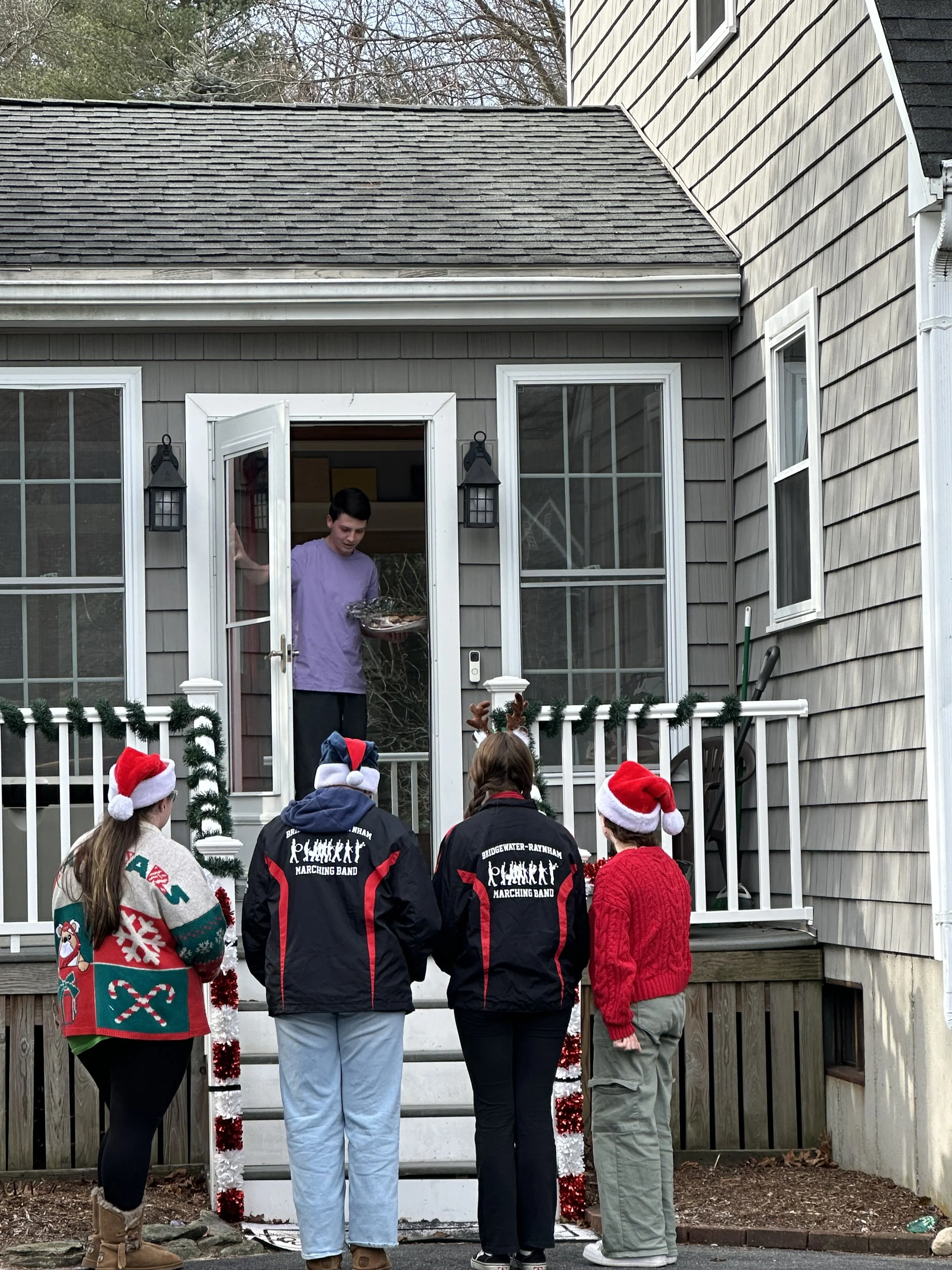 A group of people with some wearing Santa hats and sweaters, standing in front of a house with gray siding, participating in a holiday event. One person is coming out of the house holding a tray of cookies, and the house is decorated with red and white tinsel.