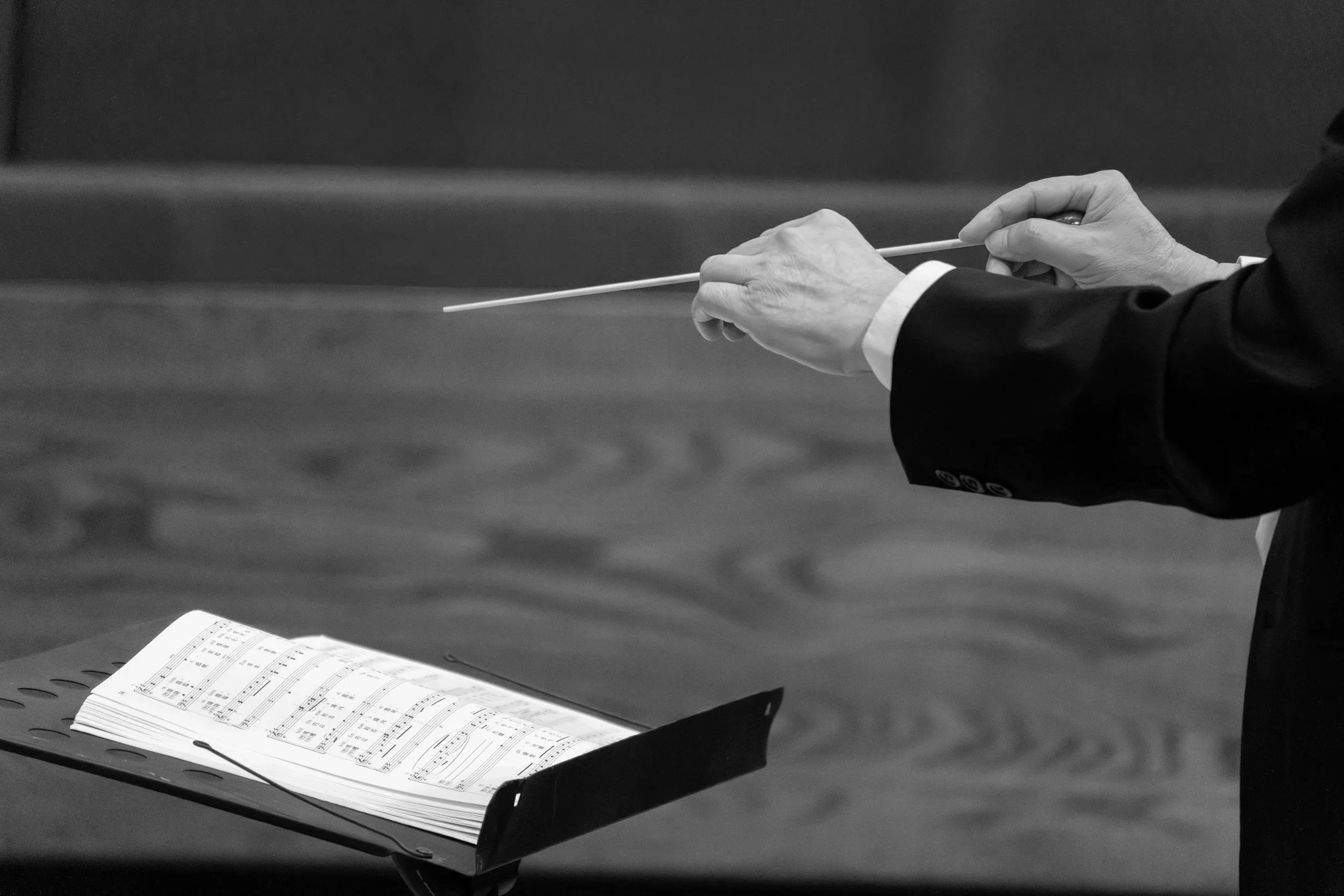 Close-up of a person's hands holding a conductor's baton, with sheet music open on a stand in front of them, in a black and white photograph.