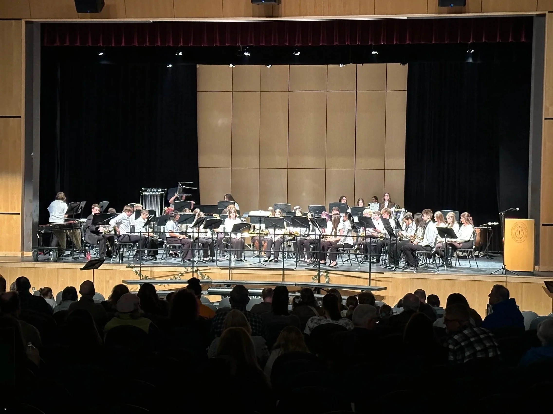 An auditorium stage with a group of people, likely a musical ensemble, seated with music stands, preparing for a performance. The audience is visible in the foreground.