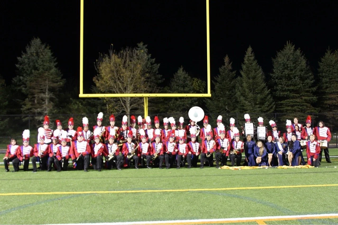 A marching band in red, white, and blue uniforms with white plumed hats, posing on a football field at night.