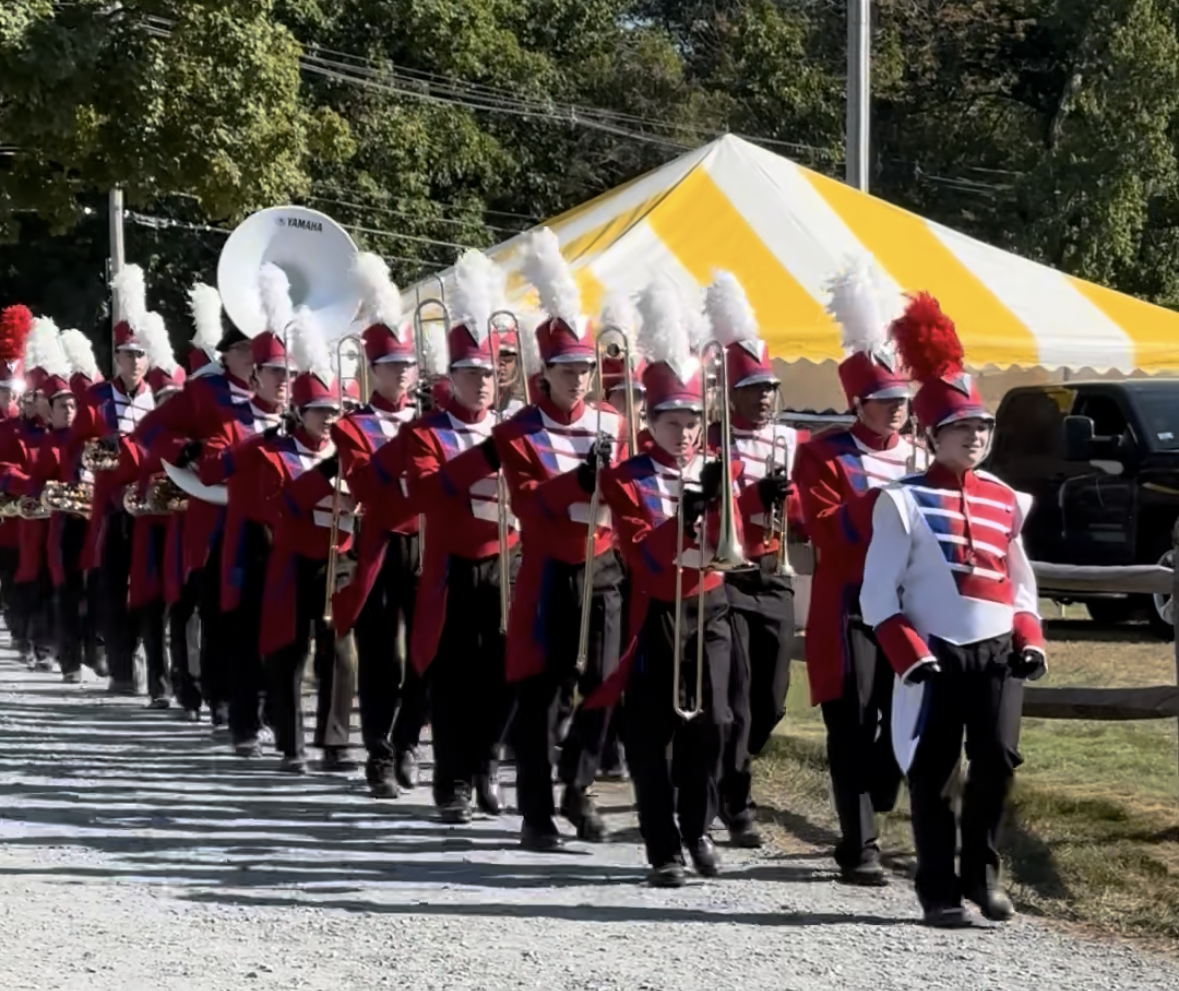 A group of young musicians playing instruments on stage during a performance, with a person conducting, and a red banner reading 'Bridgewater-Raynham Regional School District' in the background.