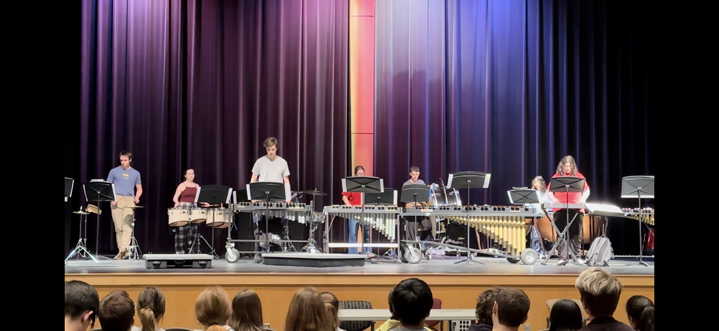 A group of musicians performing on a stage with colorful curtains, using various percussion instruments, as an audience watches.