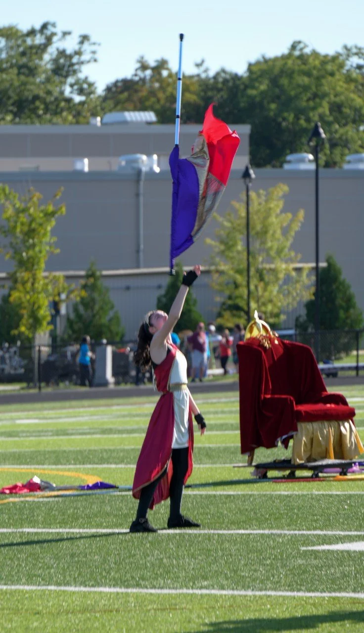 A young woman dressed in a medieval-style costume, holding a flag and standing on a football field during a performance or reenactment, with a throne draped in red fabric nearby and a group of spectators in the background.