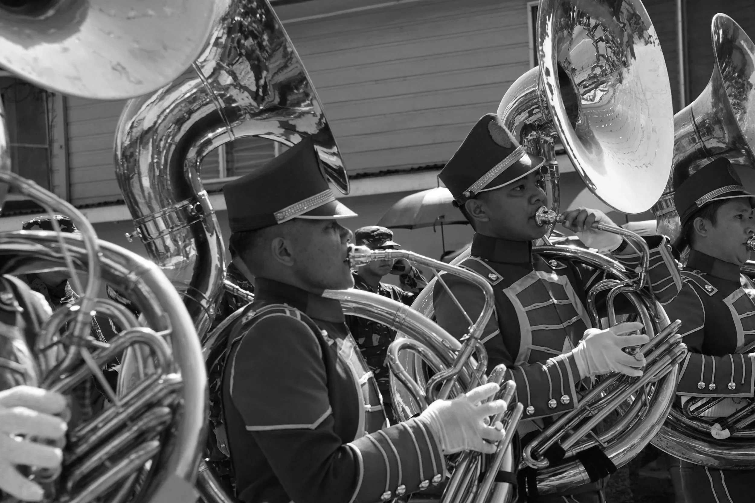 A black and white photo of marching band members playing sousaphones, wearing uniform hats and jackets, with an industrial building in the background.