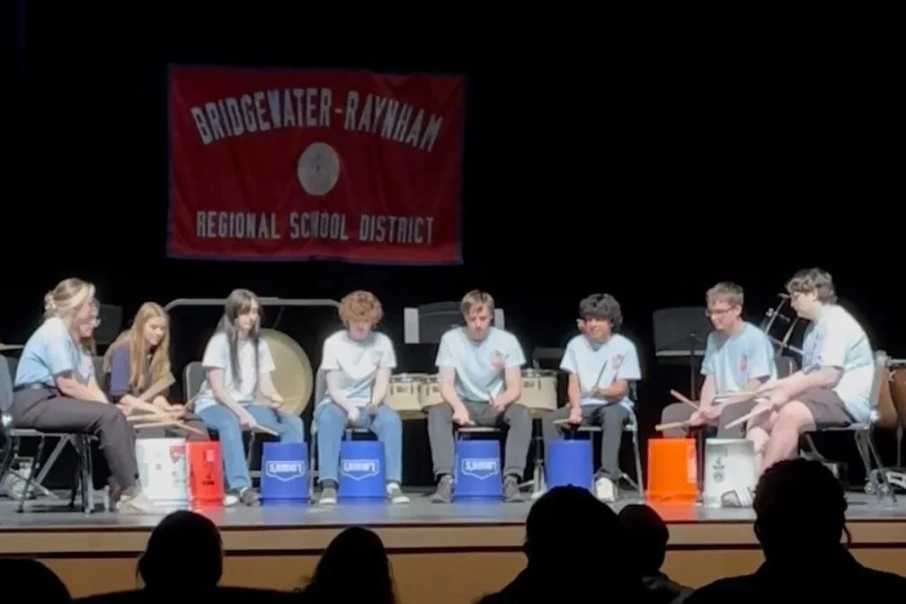 Students performing musical instruments on stage during a school event with a banner that reads "Bridgewater-Raynham Regional School District."