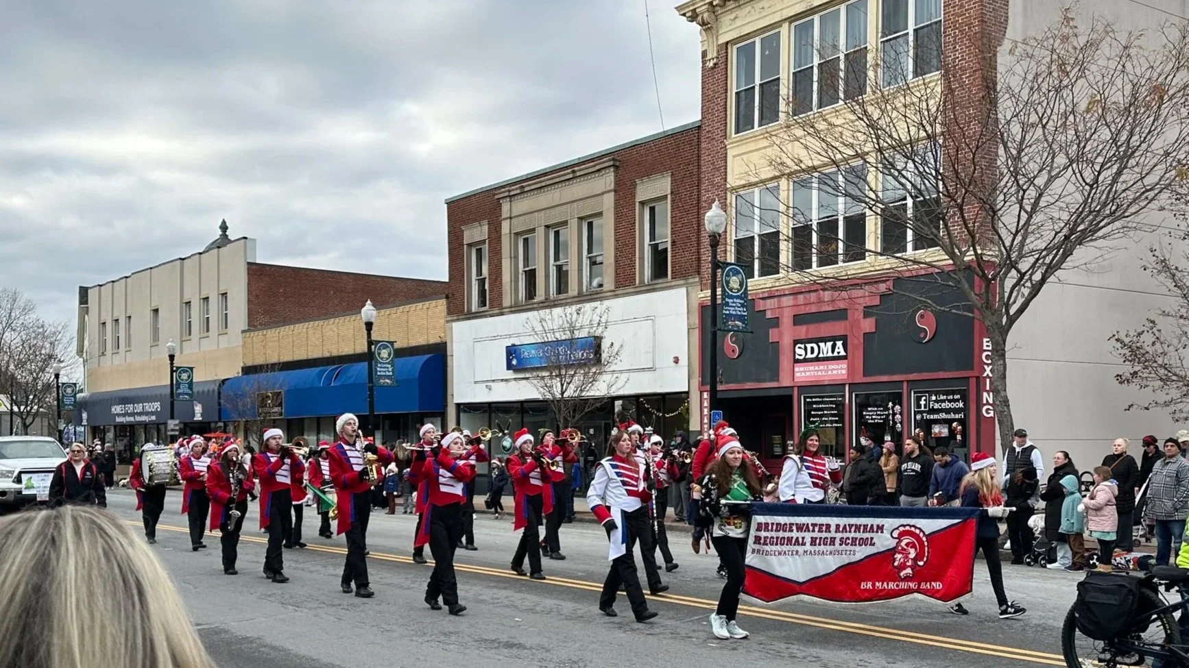 A Christmas parade on a city street featuring a marching band dressed in red, white, and black uniforms with Santa hats. The band members are playing musical instruments and holding a red banner for Bridgewater Raynham Regional High School. Spectators line the sidewalks, with storefronts and trees visible in the background.