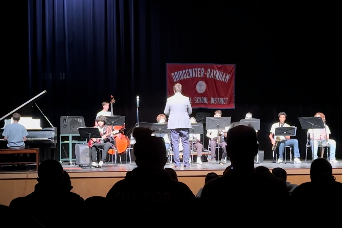 School jazz band performs on stage with conductor leading, a pianist, and various instruments including saxophones, guitar, double bass, and drums, at an event in a school auditorium.