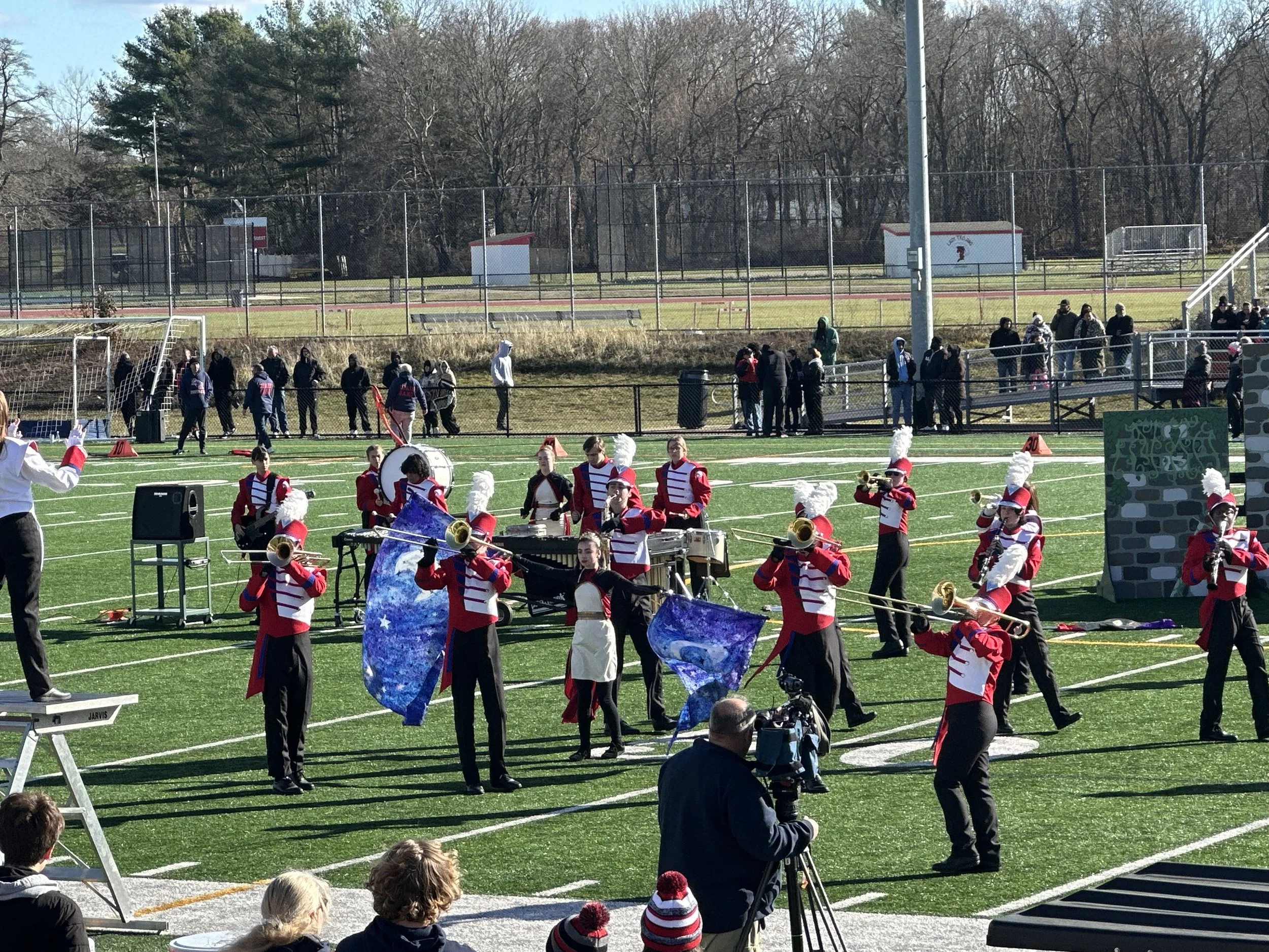 A marching band performing on a football field during a daytime event, with spectators and a fence in the background.