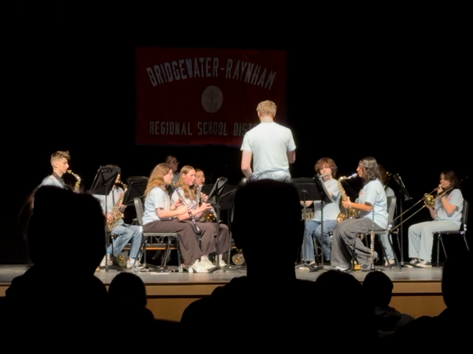 Young students performing on stage in a school band, directed by a conductor, at a regional school district event.