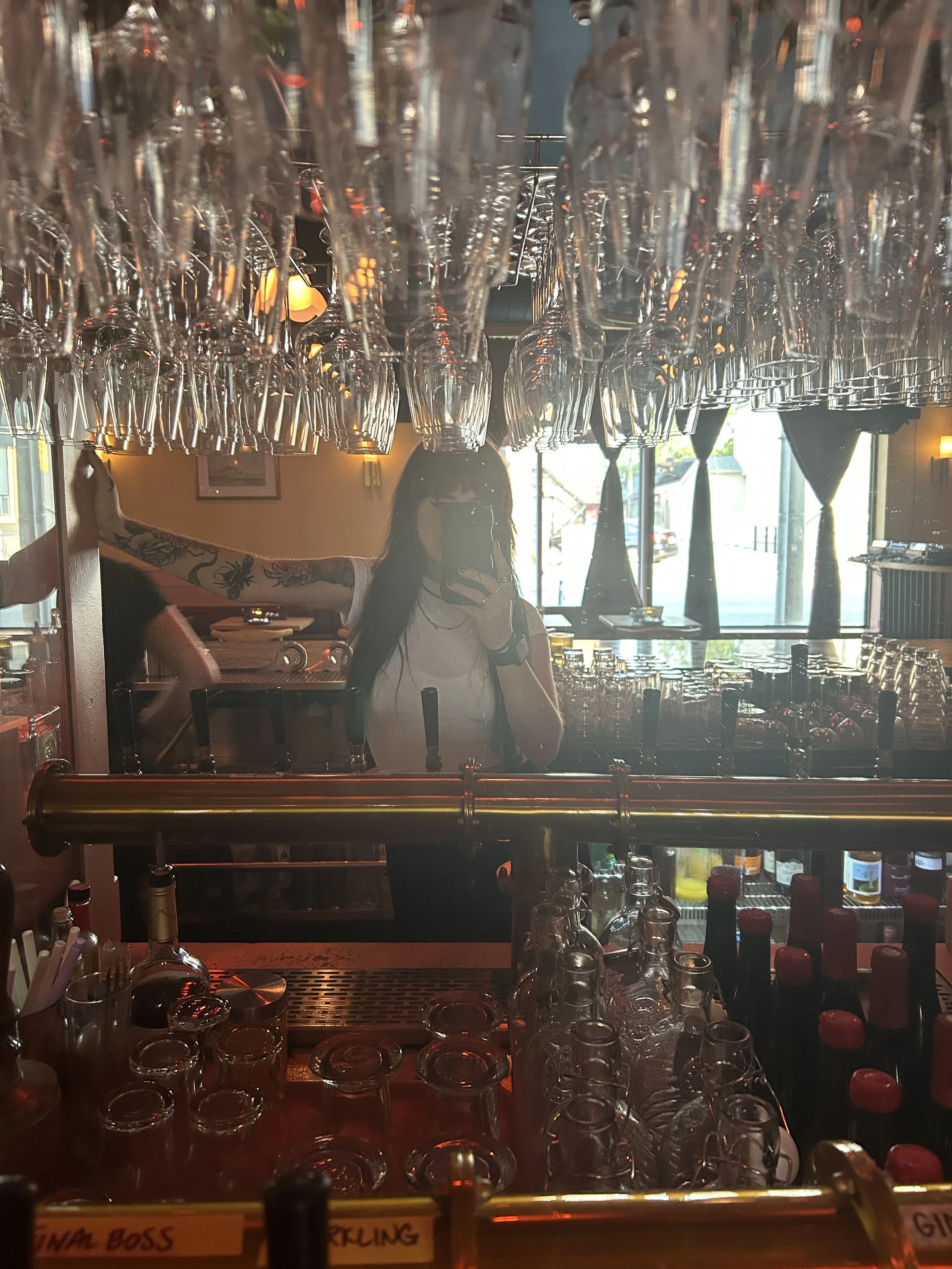 A mirror behind a bar with many upside-down wine glasses hanging from a rack above a counter. The bartender is reflected in the mirror, taking a photo with her phone. The bar is stocked with bottles and bar tools, and sunlight is coming through the window.