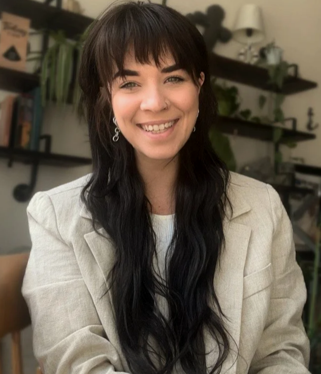 Smiling woman with long dark wavy hair, wearing a beige blazer and earrings, sitting indoors with shelves and plants in the background.