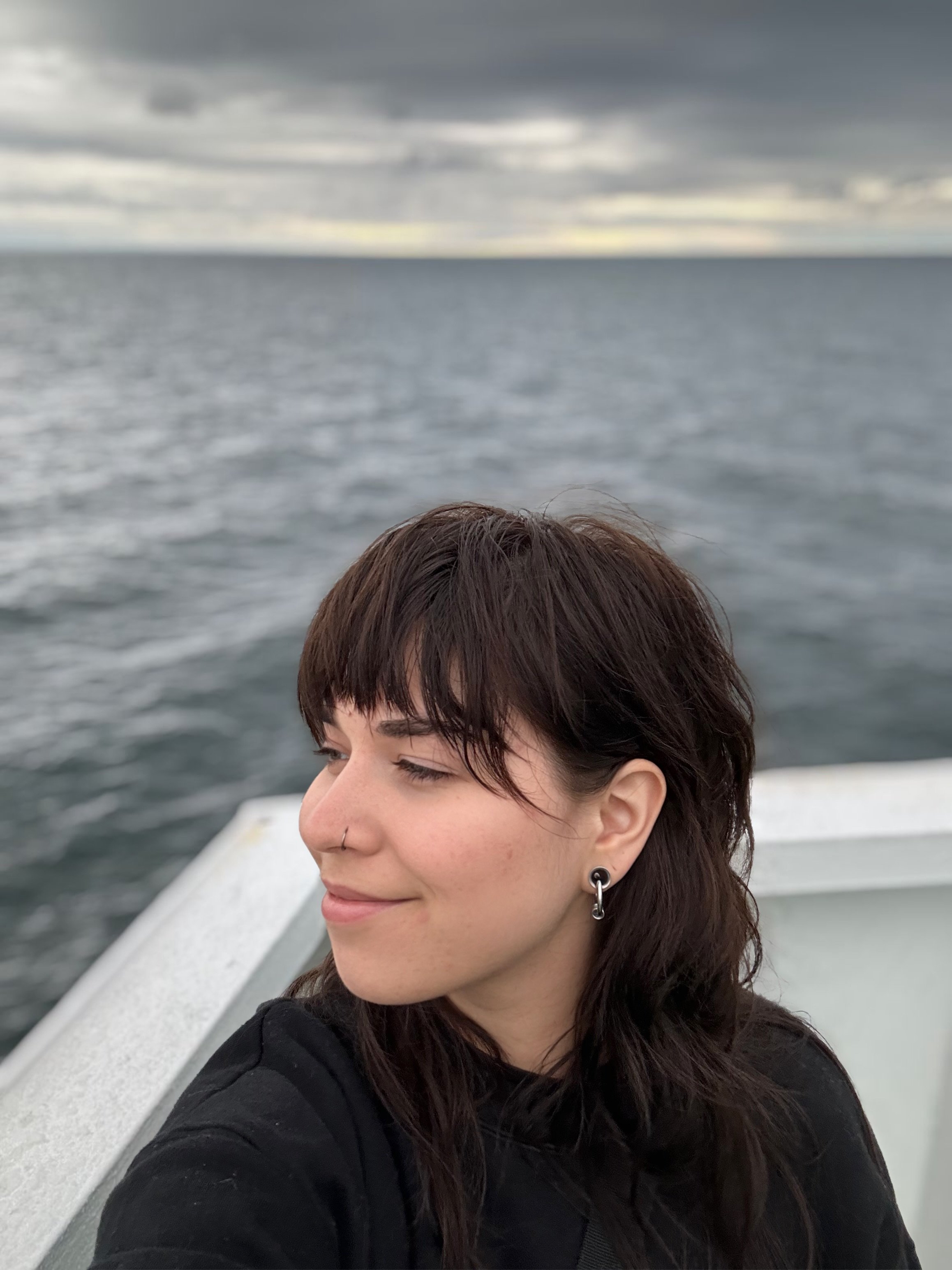 A young woman with long dark hair, closed eyes, earrings, and a nose piercing, on a boat or ferry with water and cloudy sky in the background.