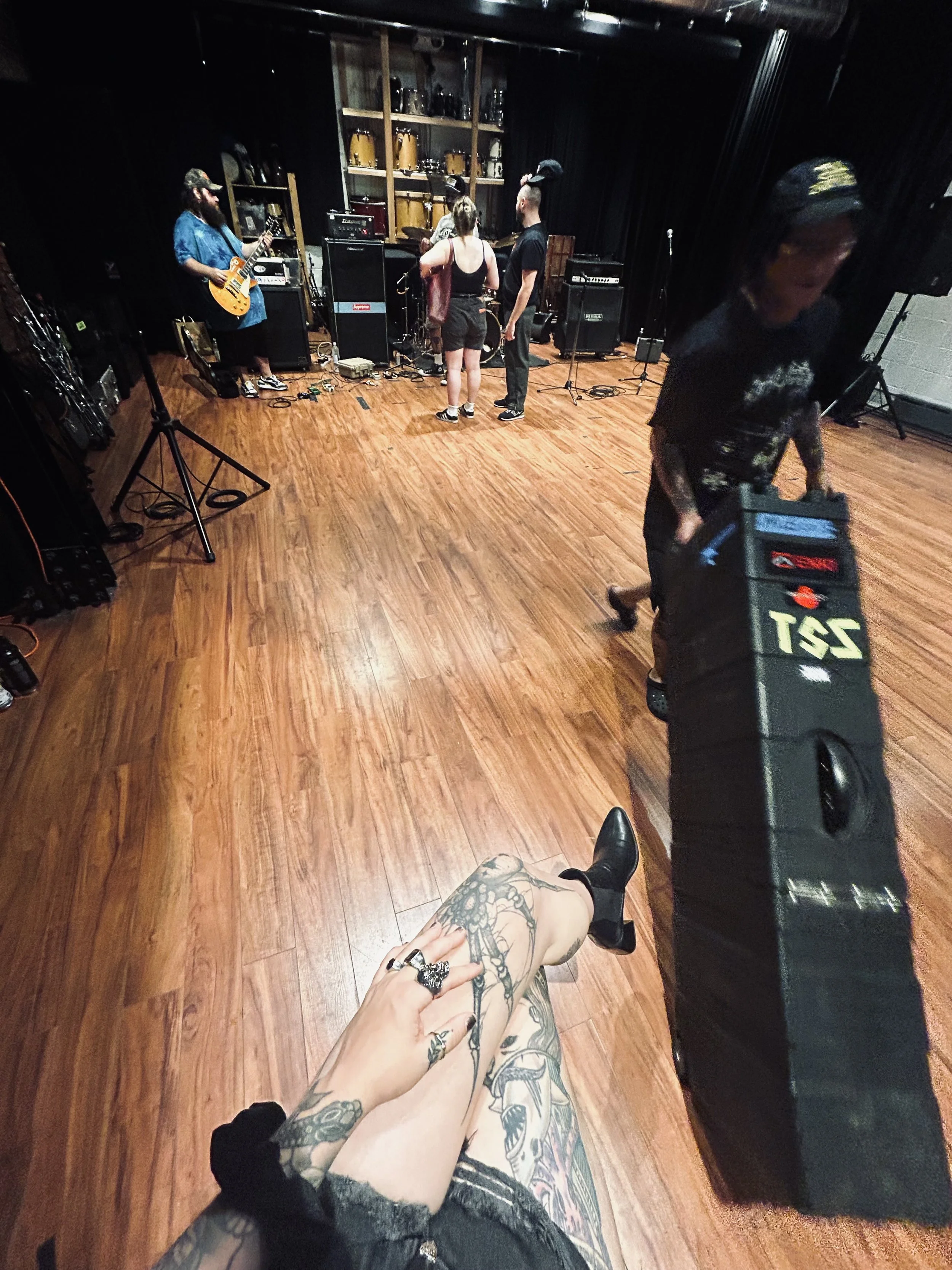 A person with tattooed arms and legs is sitting cross-legged on the floor, holding their leg in their hand, with visible rings on their fingers, and wearing black boots, in a music rehearsal or recording studio. The studio has wooden flooring, black curtains, and shelves with musical instruments and equipment. In the background, there are band members setting up or adjusting their instruments, including a guitarist and a person with bunny ears headband.