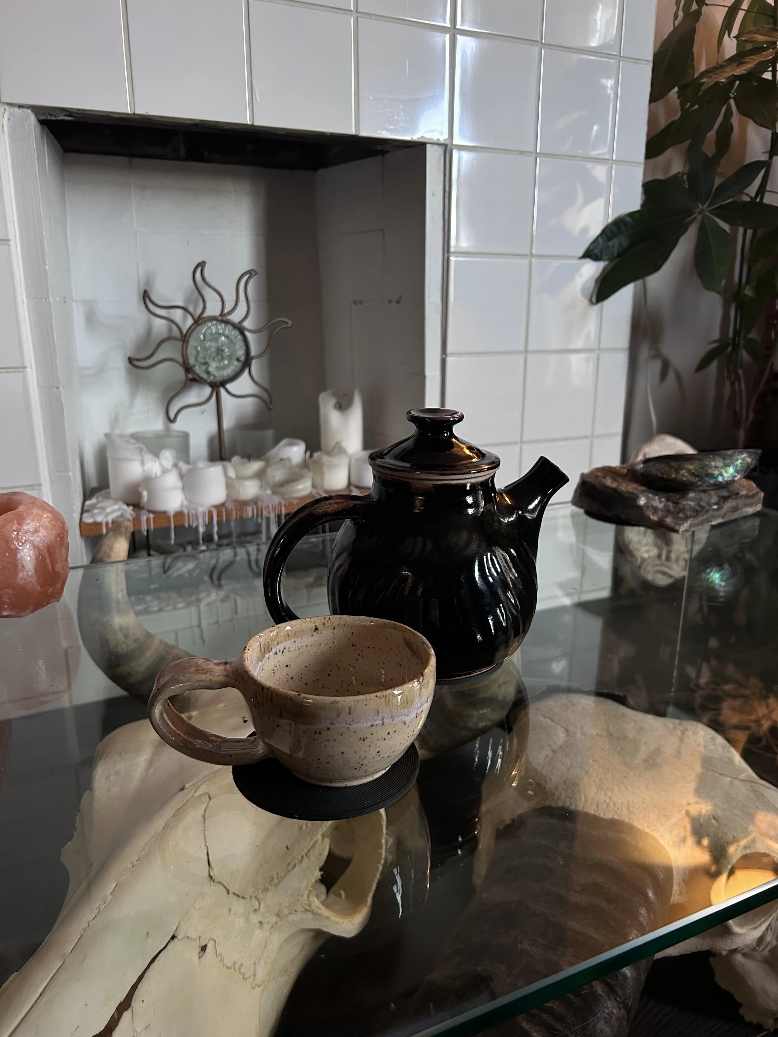 Close-up of various decorative items on a glass table, including a black ceramic teapot, a beige ceramic mug, a skull-shaped object, and other minerals and shells, with a background of white tiled wall, candles, and plants.