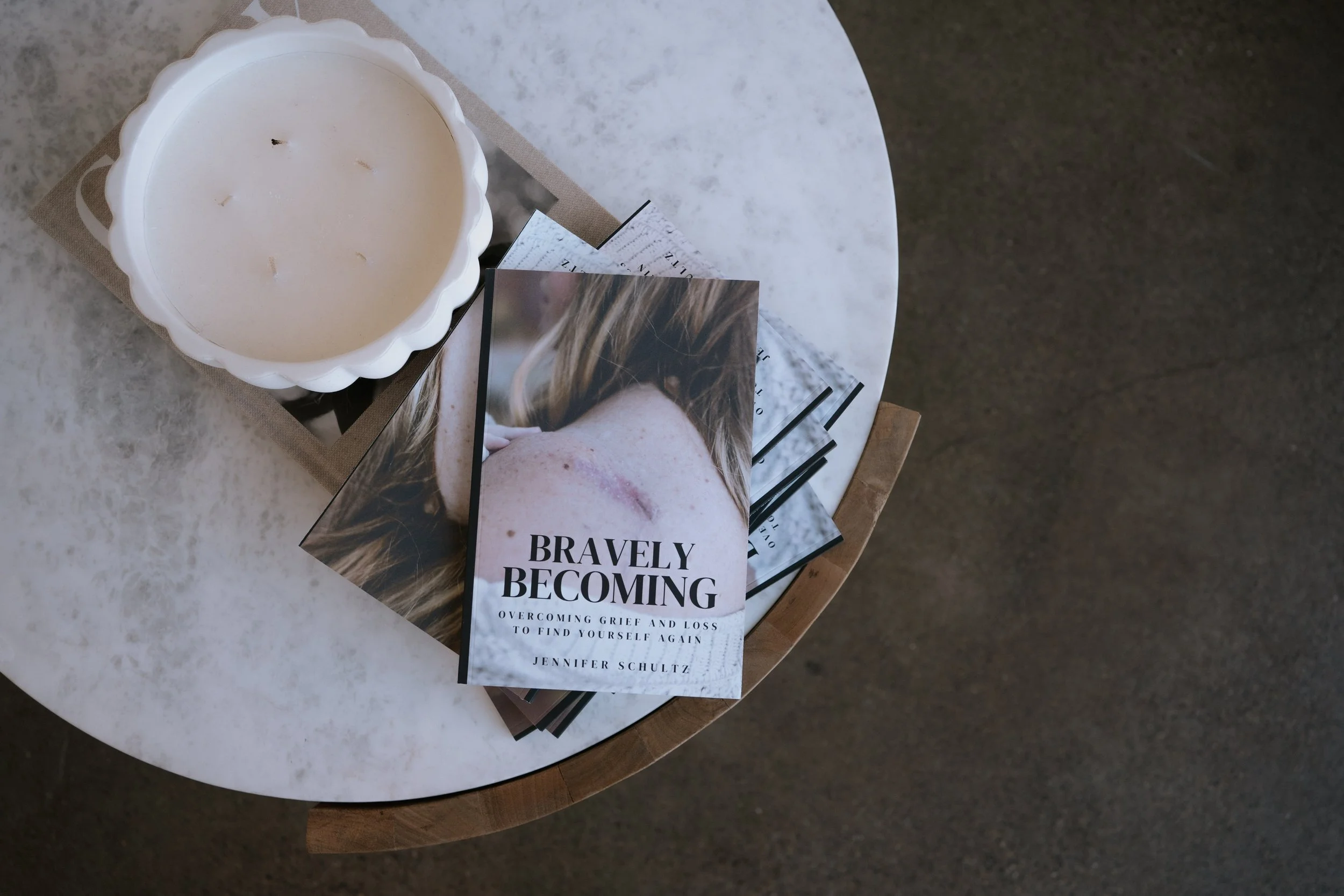 A white candle in a scalloped ceramic holder on a small round table with magazines and a stack of books titled 'Bravely Becoming,' focusing on overcoming grief and loss.