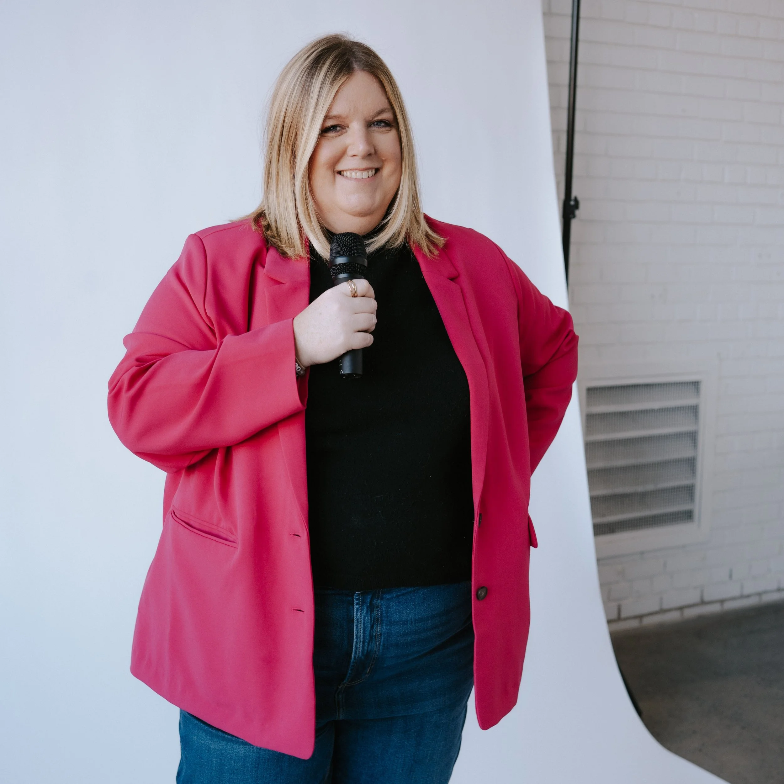 Jenn Patrice Schultz, a woman with blonde hair wearing a pink blazer and blue jeans, smiling and holding a microphone. She is standing indoors against a white wall with a brick wall visible on the right.
