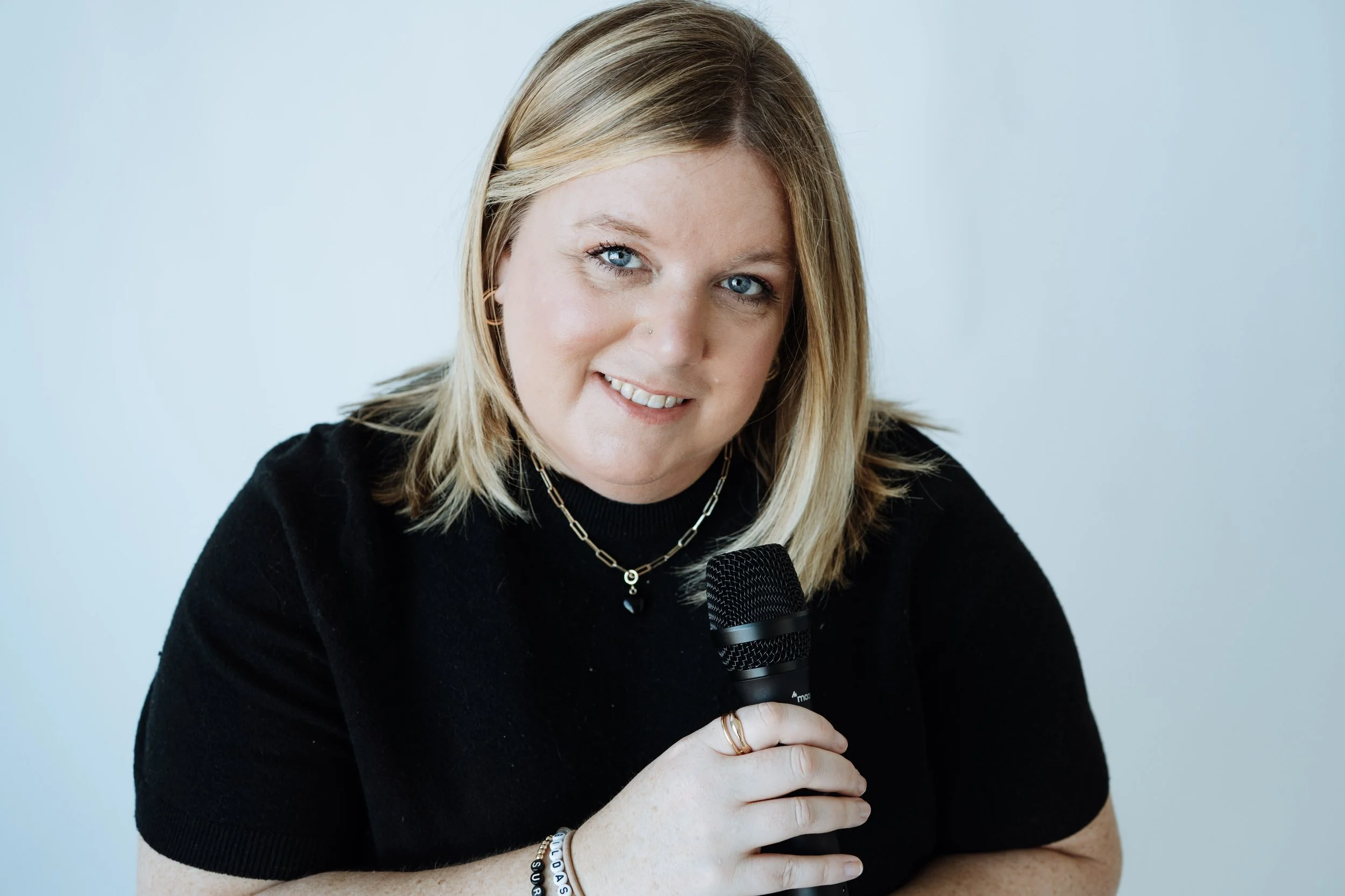 Jenn Patrice Schultz, a woman with blonde hair, blue eyes, and fair skin, holding a microphone and smiling, against a light blue background.