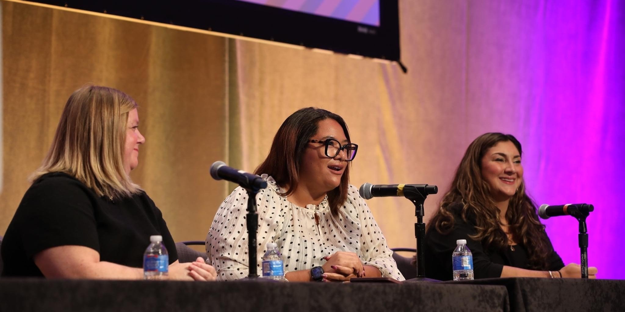 Three women, one of whom is Jenn Patrice Schultz, sitting at a panel discussion, each with a microphone and water bottles in front of them, against a background with purple lighting.