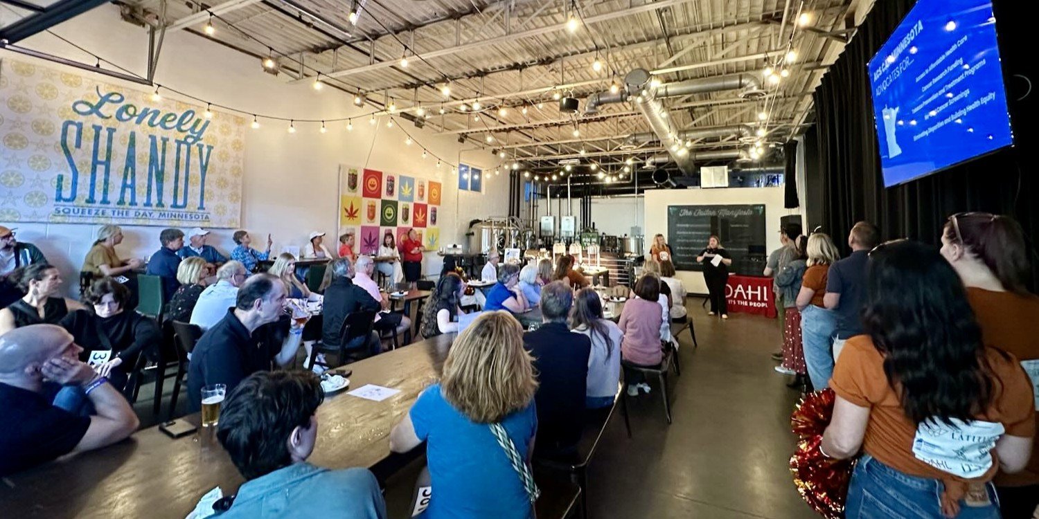 People attending an American Cancer Society event in a large indoor venue with industrial ceiling and string lights Some people are seated at tables, others stand along the sides, and Jenn Patrice Schultz is the mission speaker at the front.