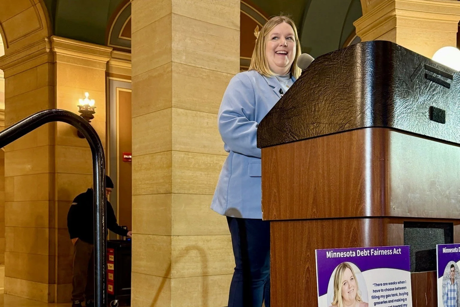 Jenn Patrice Schultz, a woman with blond hair wearing a light blue blazer, standing at a podium, speaking or presenting, at the Minnesota State Capitol.