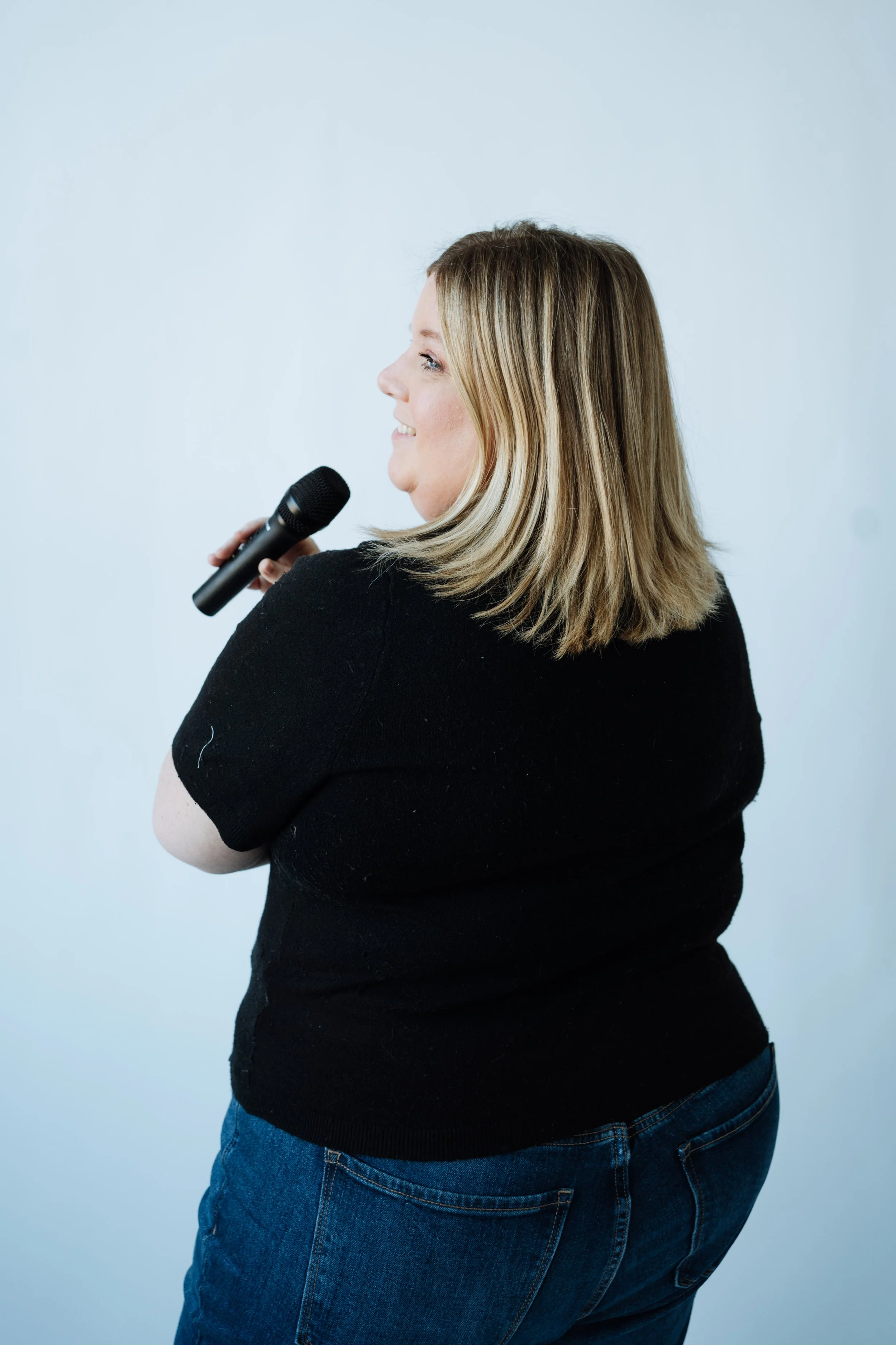 Jenn Patrice Schultz, a woman with shoulder-length blonde hair smiling and holding a microphone against a plain light blue background.