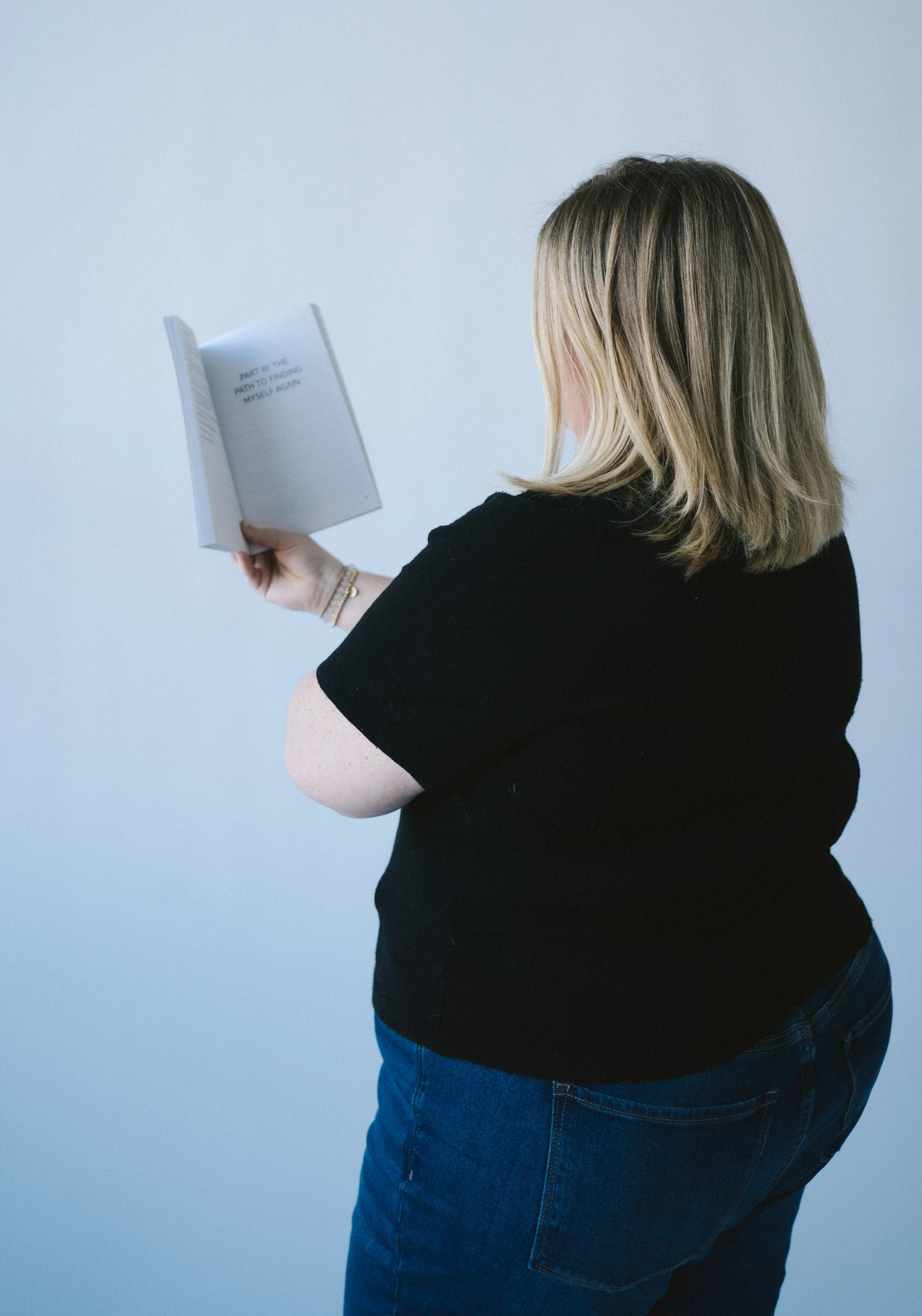Back view of Jennifer Schultz reading her book titled "Bravely Becoming," against a plain light blue background.